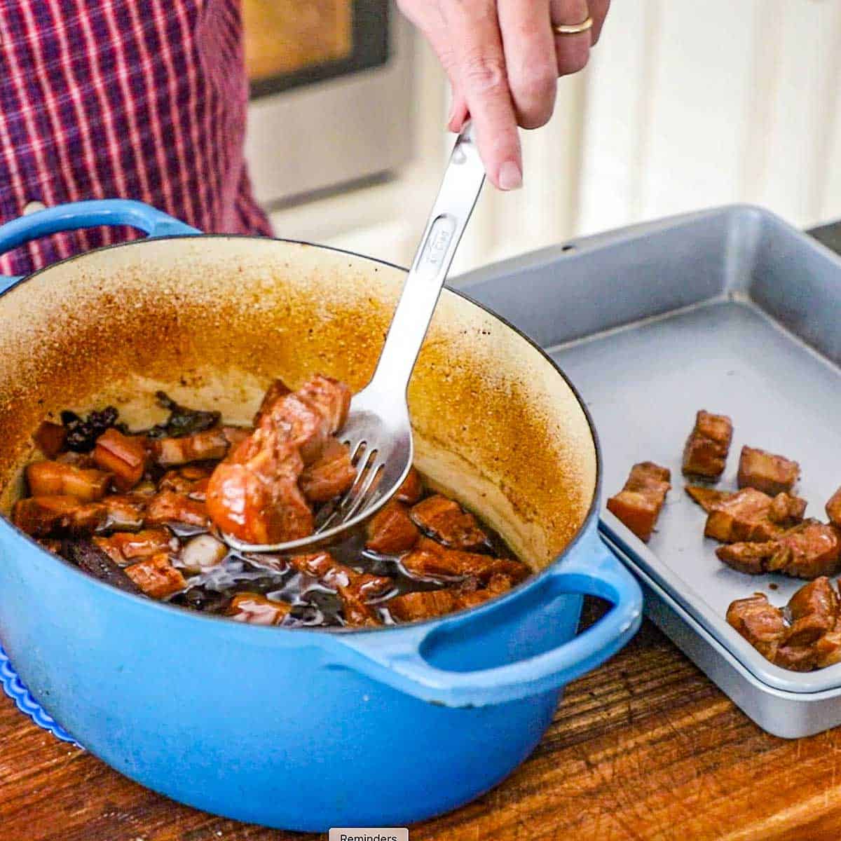 A person using a slotted spoon to transfer braised pork belly chunks from a brown sauce in a Dutch oven into a metal pan nearby.