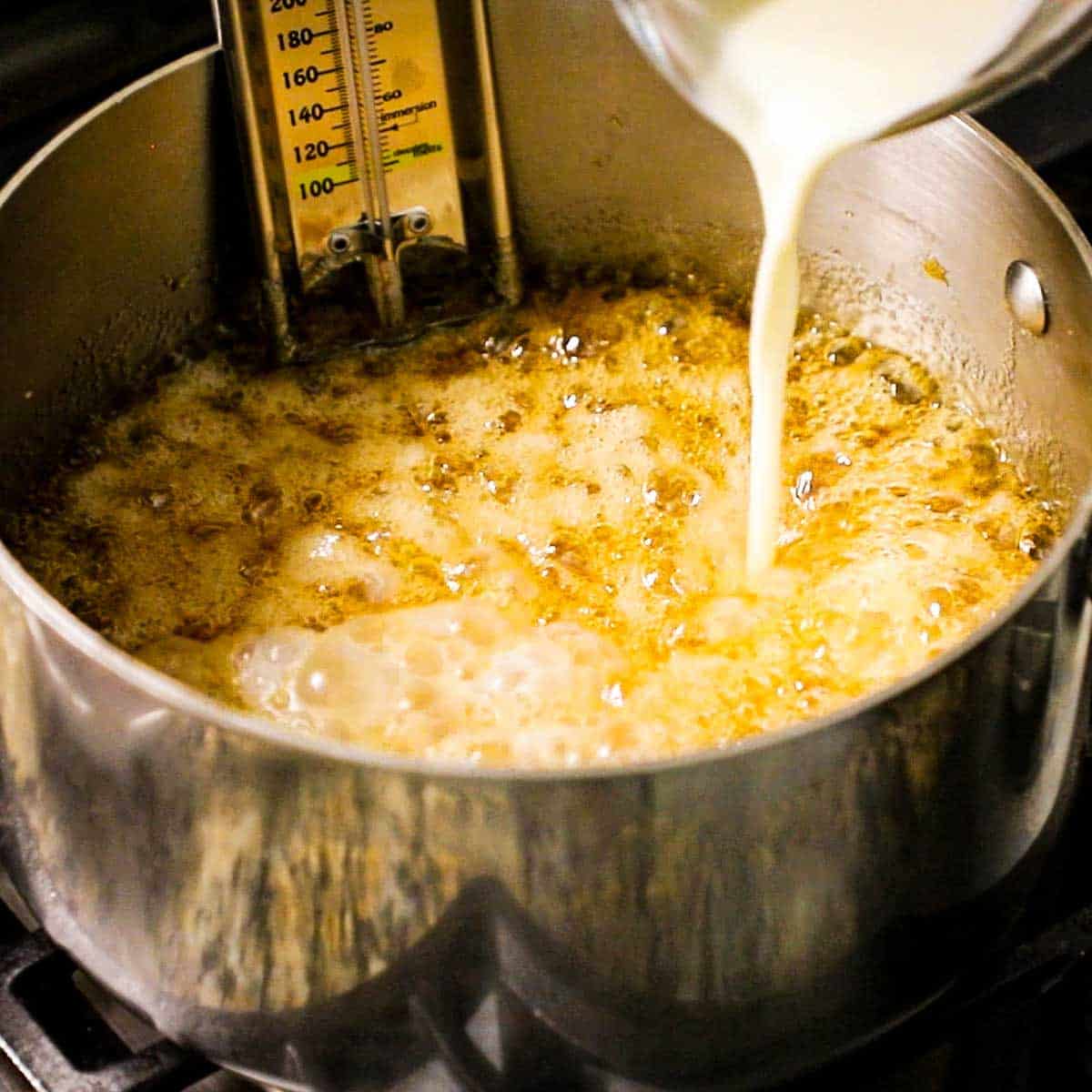 A person pouring warm heavy cram from a glass measuring cup into a very hot mixture of amber-colored melted sugar with honey.