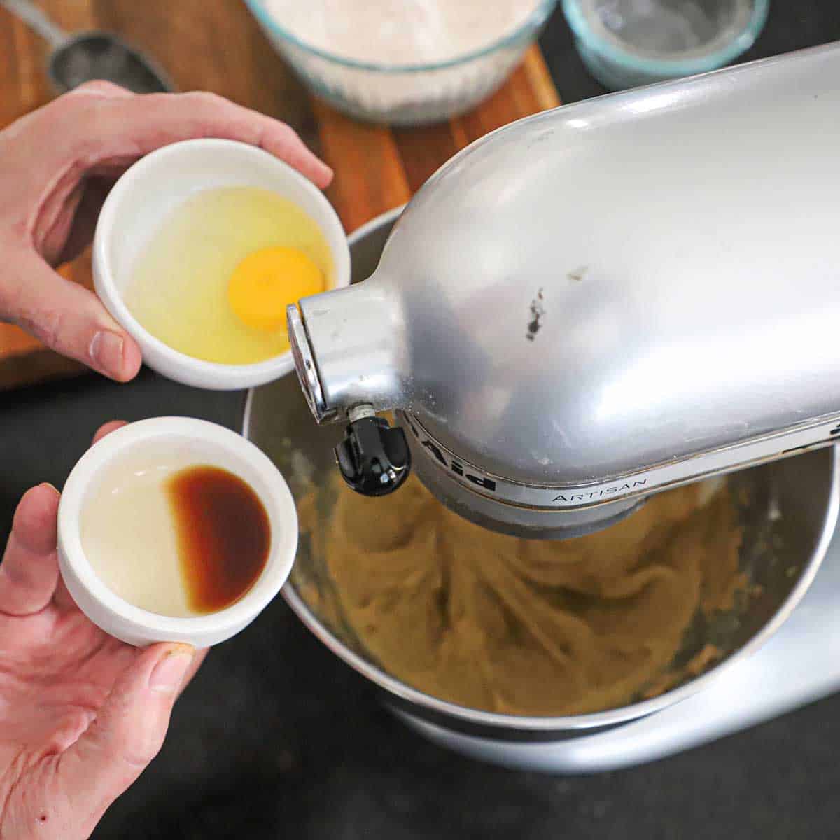 A person holding a small bowl filled with an egg in one hand and another bowl of vanilla extract in his other hand both over a stand mixer that is filled with a pumpkin coffee cake batter.