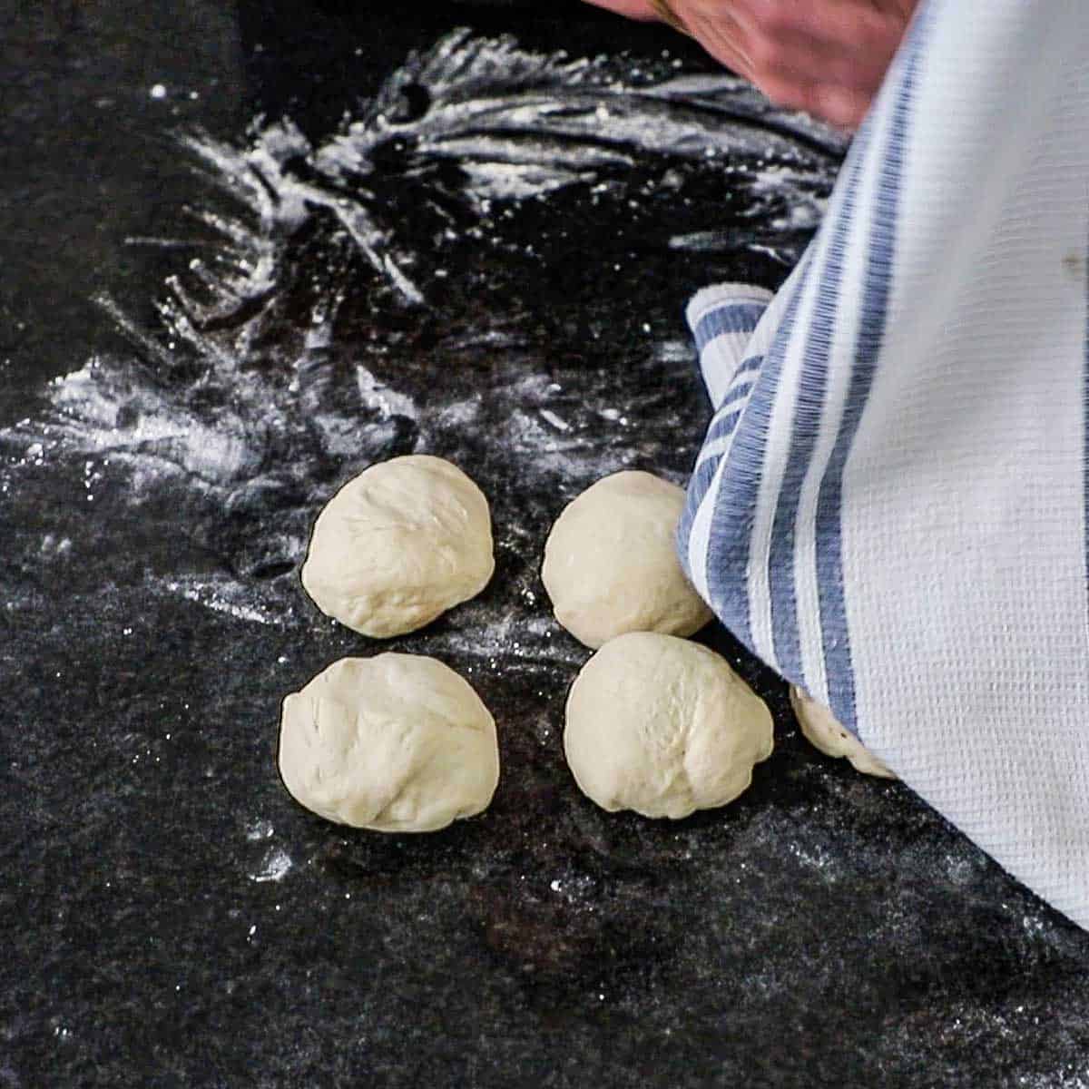 A person draping a kitchen towel over balls of homemade telera dough on a lightly floured black granite counter.