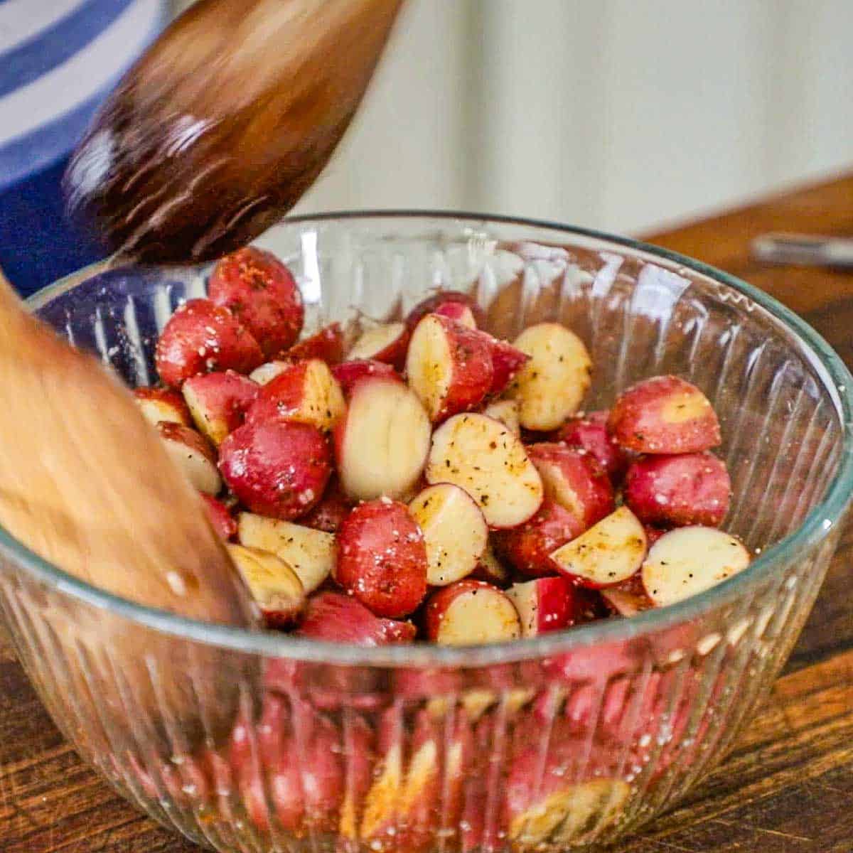 A person using two large wooden spoons to toss halved red baby potatoes in a large glass bowl along with olive oil and a seasoning mixture.