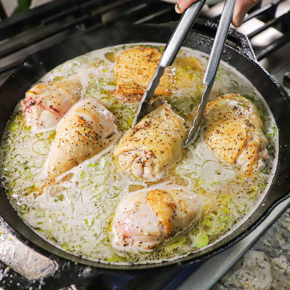 A person using a pair of tongs to place a partially cooked skin-on chicken thigh in a large cast-iron skillet that is filled with a simmering sauce along with other pieces of chicken.