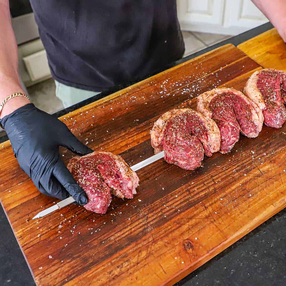 A person sliding the last of four folded seasoned picanha steaks onto a large skewer that is resting on a large wooden cutting boards.