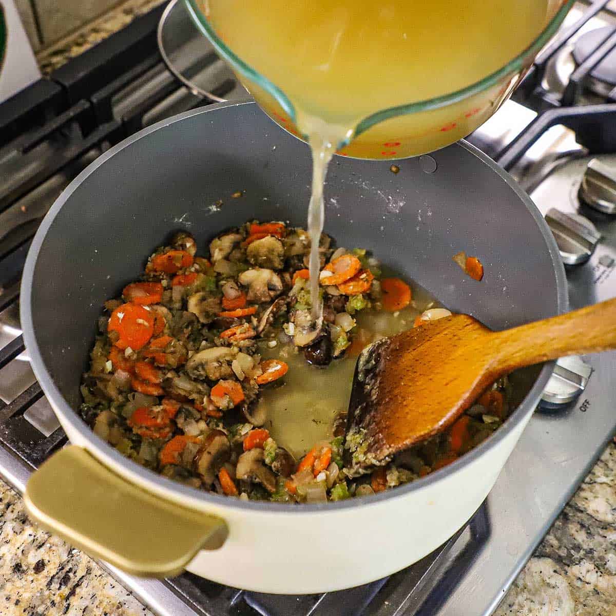 A person pouring chicken stock from a large glass measuring cup into a large pot filled with sautéed vegetables that have been coated with all-purpose flour.