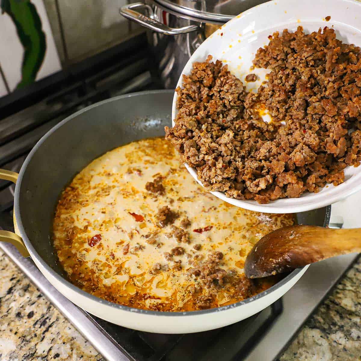 A person transferring cooked crumbled Italian sausage from a white bowl into a large skillet filled with a simmering Tuscan cream sauce.