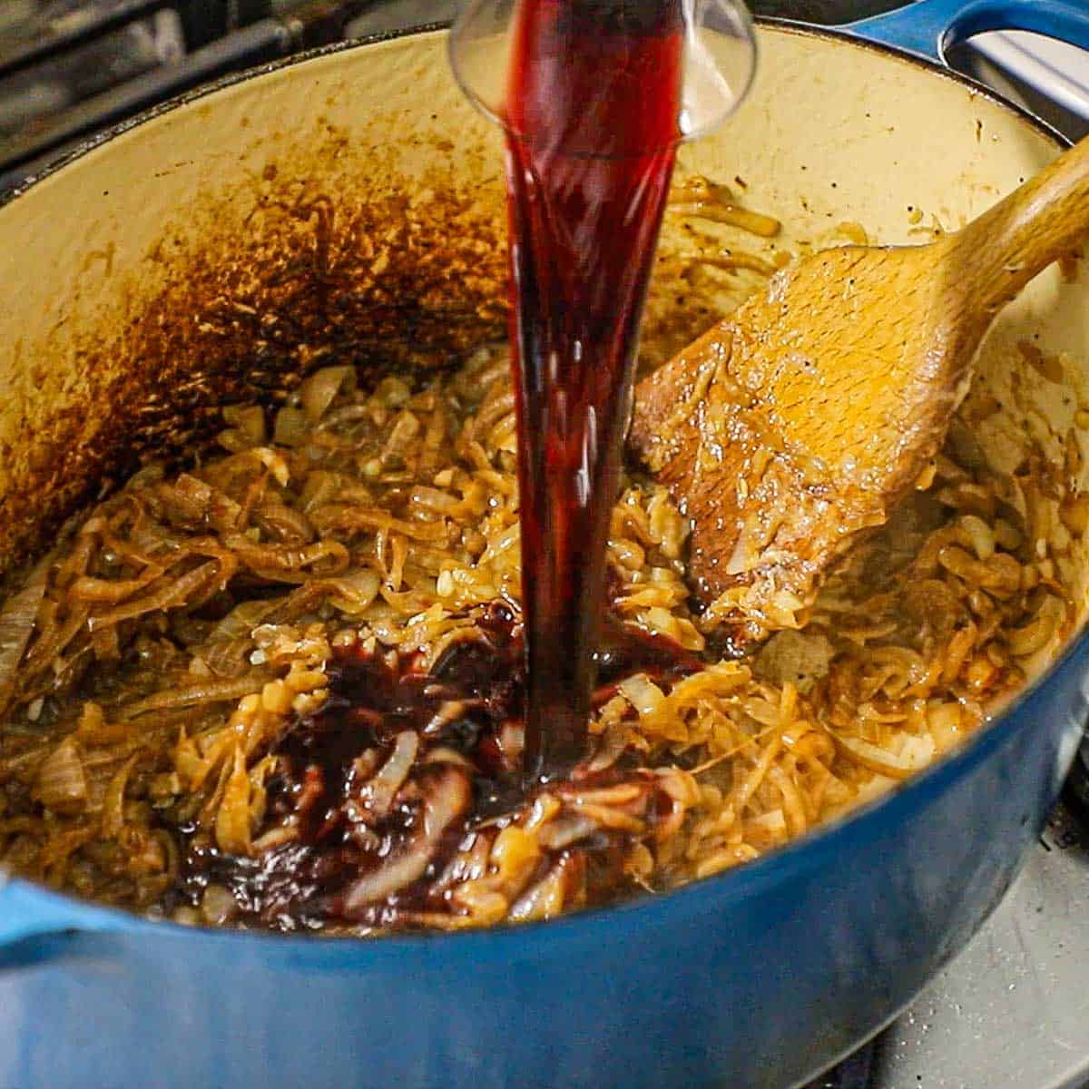 A person pouring red wine from a small carafe into a large oval Dutch oven that is filled with simmering caramelized onions.