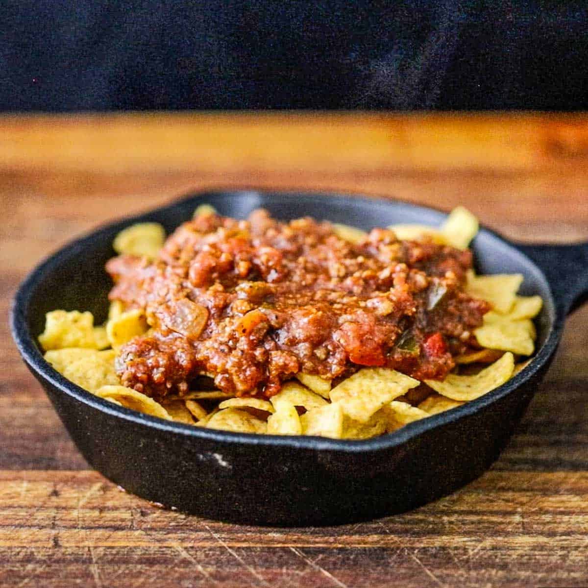 A straight-on view of a small cast-iron skillet filled with corn chips topped with steaming beef chili.