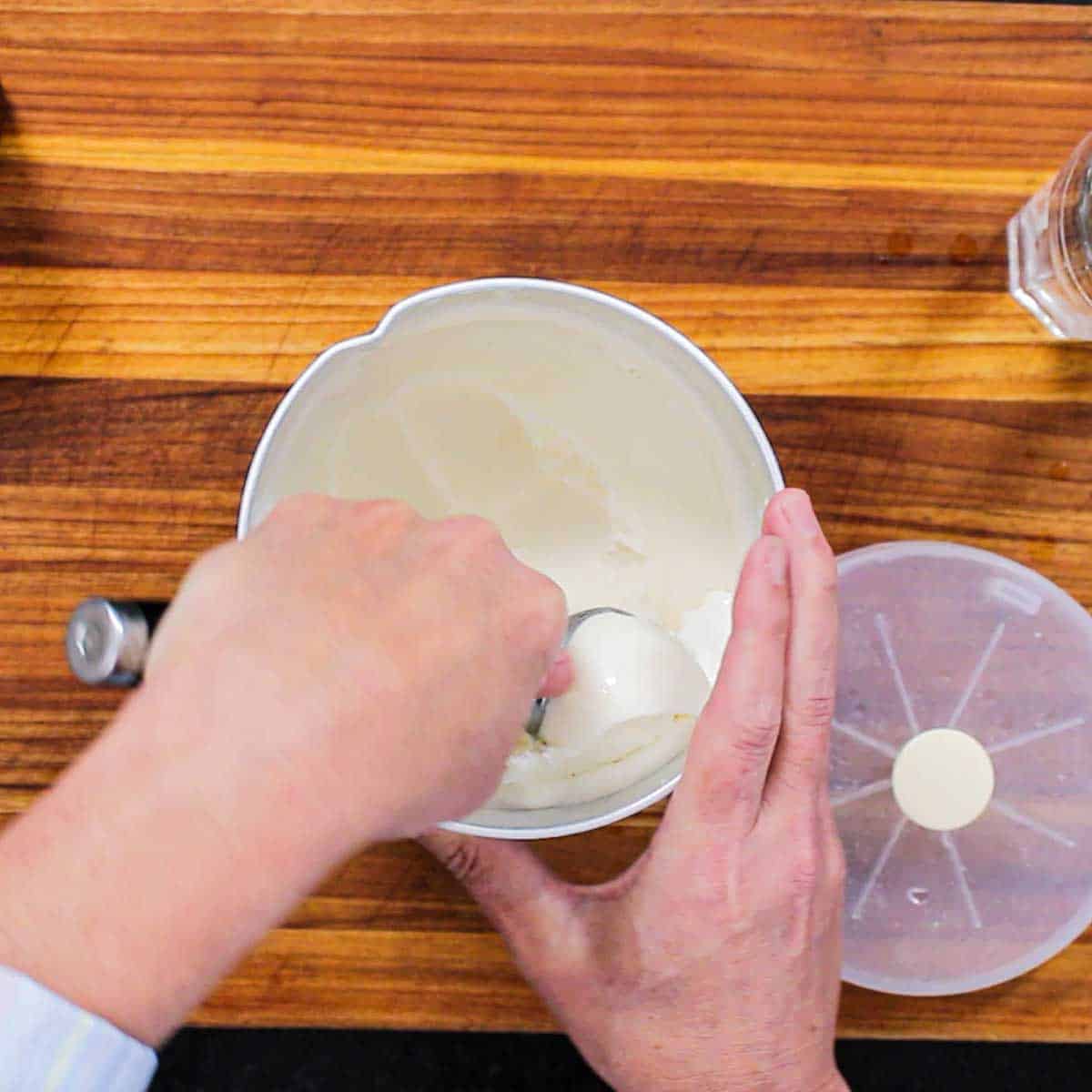 A person using an ice cream scooper to form a ball of homemade ice cream from a metal ice cream container on a large wooden cutting board.