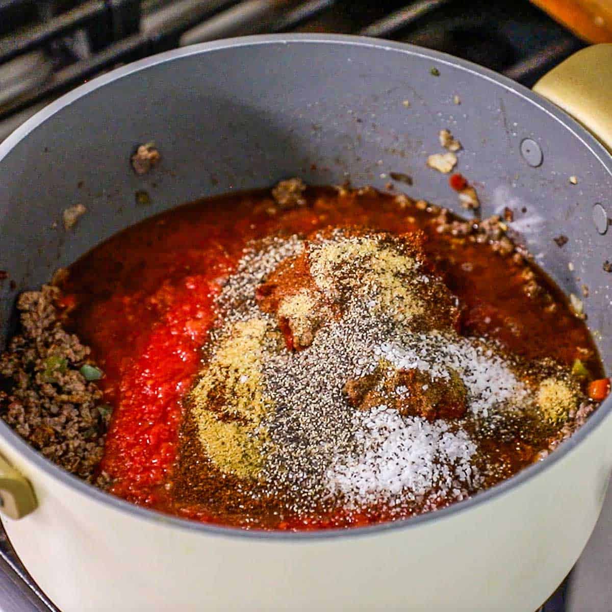 Large pot filled with cooked ground beef along with chopped tomatoes, beef broth, tomato paste, chili powder, and other seasonings for beef chili over a gas stove.