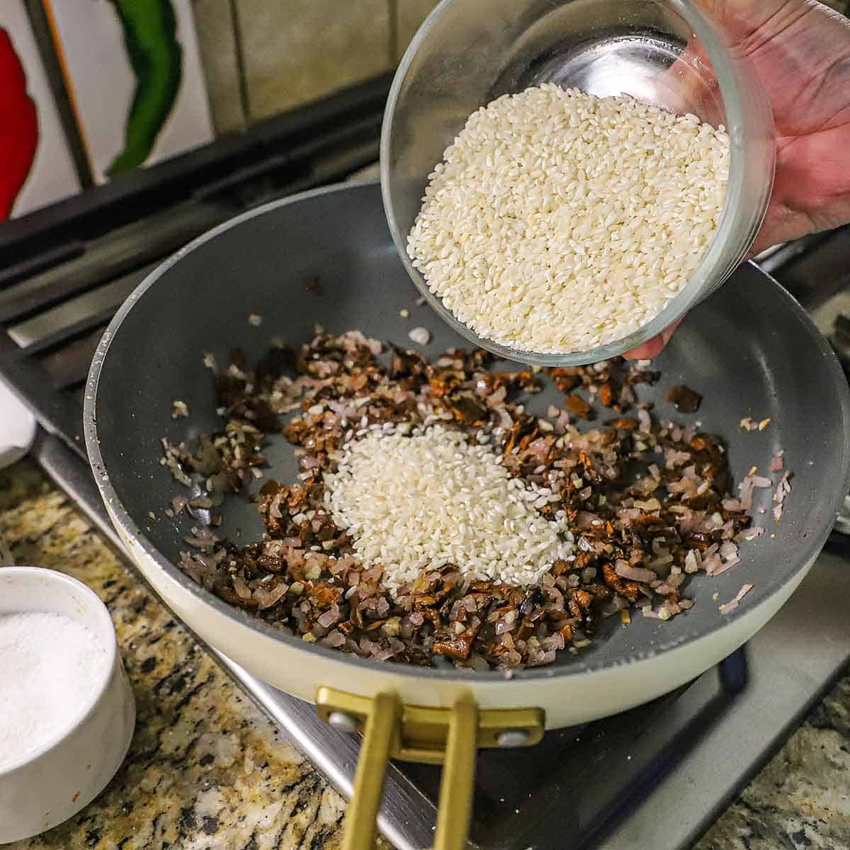 A person transferring arborio rice from a glass bowl into a large non-stick skillet filled with saut&eacute;ed shallots, garlic, and chopped porcini mushrooms.