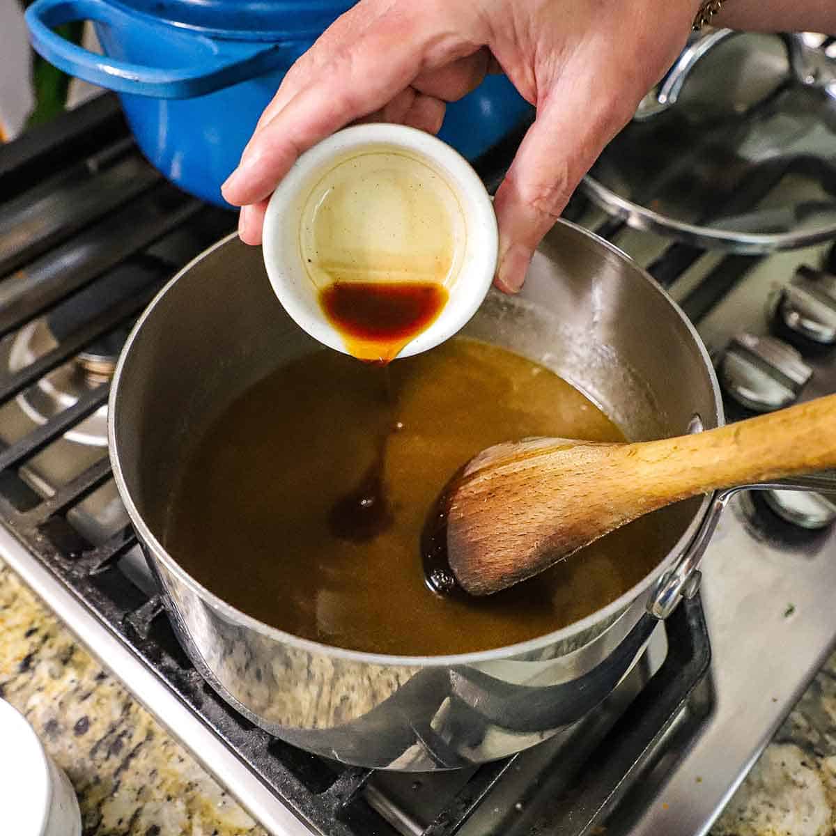 A person pouring vanilla extract from a small white bowl into a saucepan on a gas stove filled with brown sugar that has dissolved in simmering melted butter.
