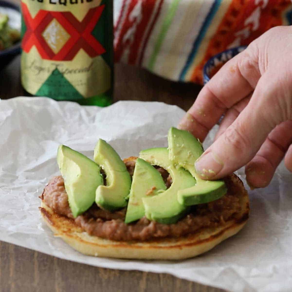 A person placing slices of avocado over a thin layer of refried beans that have been spread across the bottom half of a toasted telera bun.