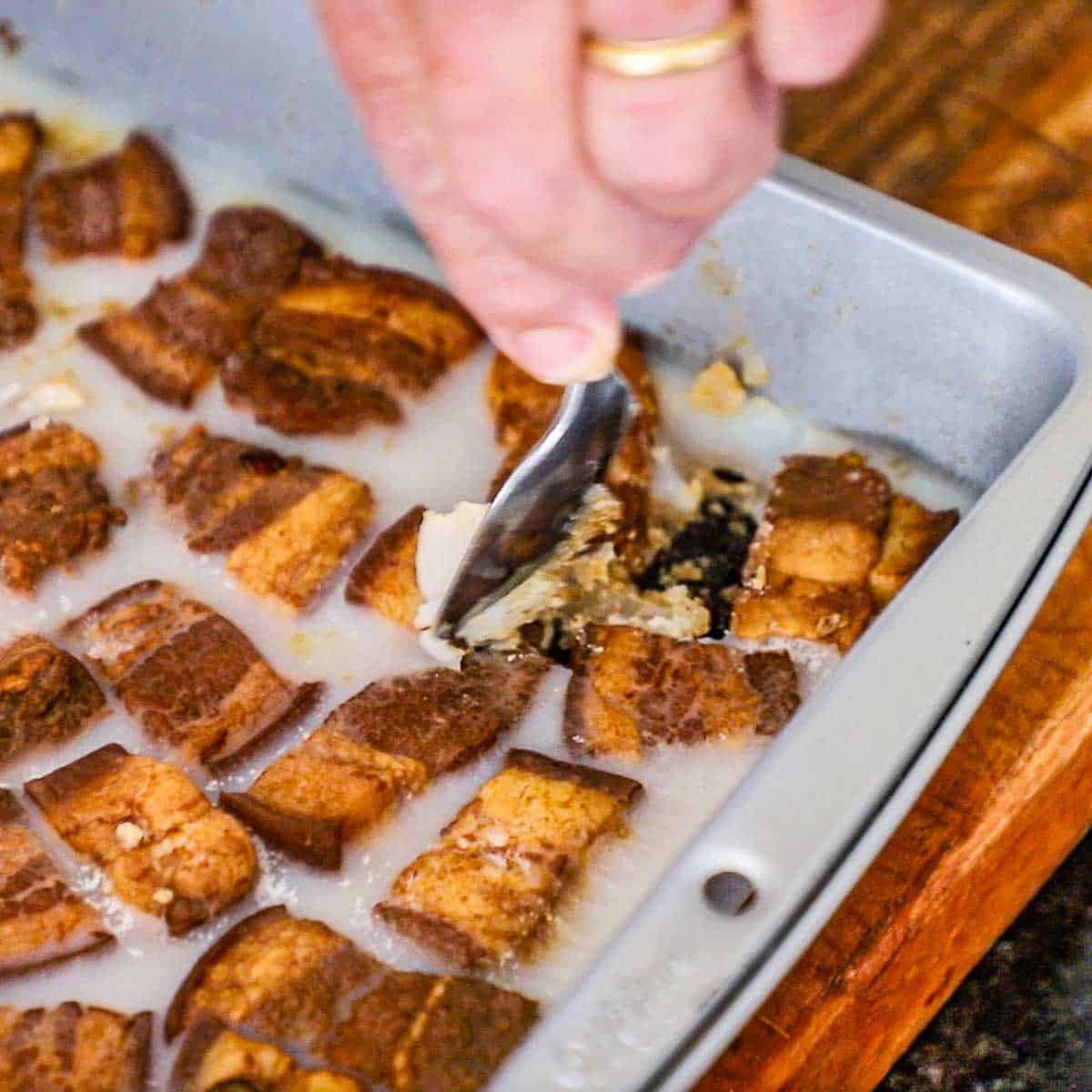 A person using a small spoon to scrape off solidified fat that has formed along the sides of chilled braised pork chunks in a liquid in a metal pan on wooden cutting board.