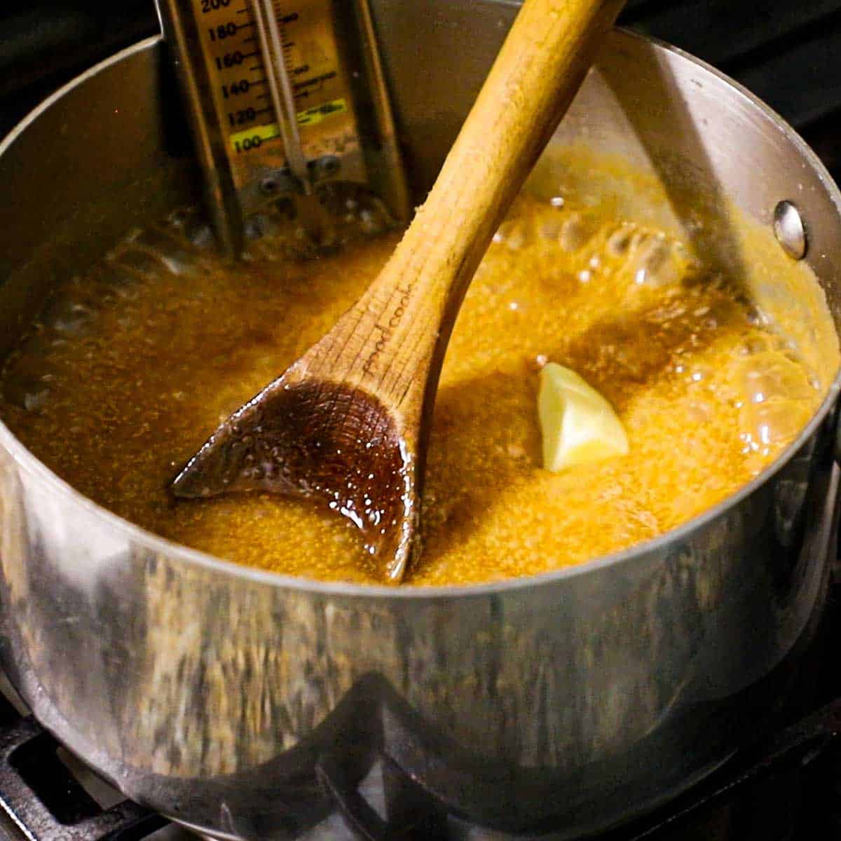 A wooden spoon being used to stir melting butter in a saucepan that is filled with hot caramel sauce.