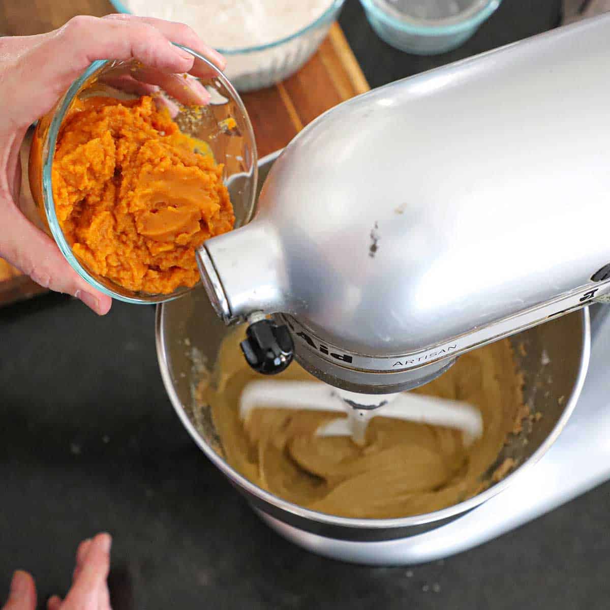 A person transferring pumpkin purée from a glass bowl into a stand mixer that is beating a pumpkin spice coffee cake batter.