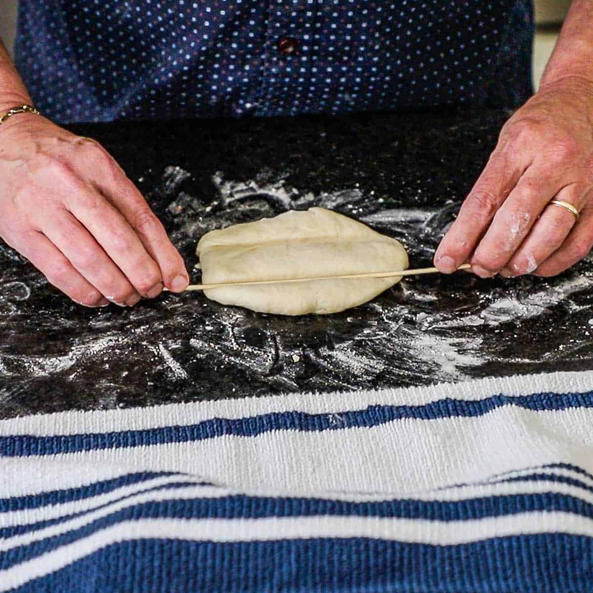 A person using a thin wooden skewer to created an end-to-end indention in an oval piece of bread dough on a lightly floured kitchen counter.