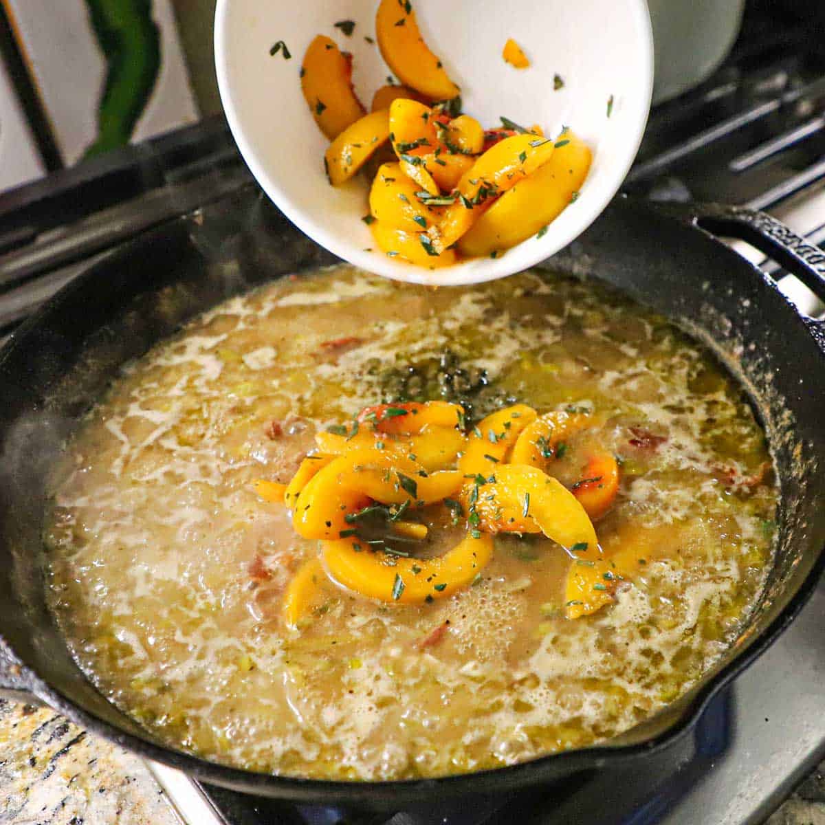 A person transferring marinated peaches from a white bowl into a cast-iron skillet that is filled with a simmering broth and leeks sauce.