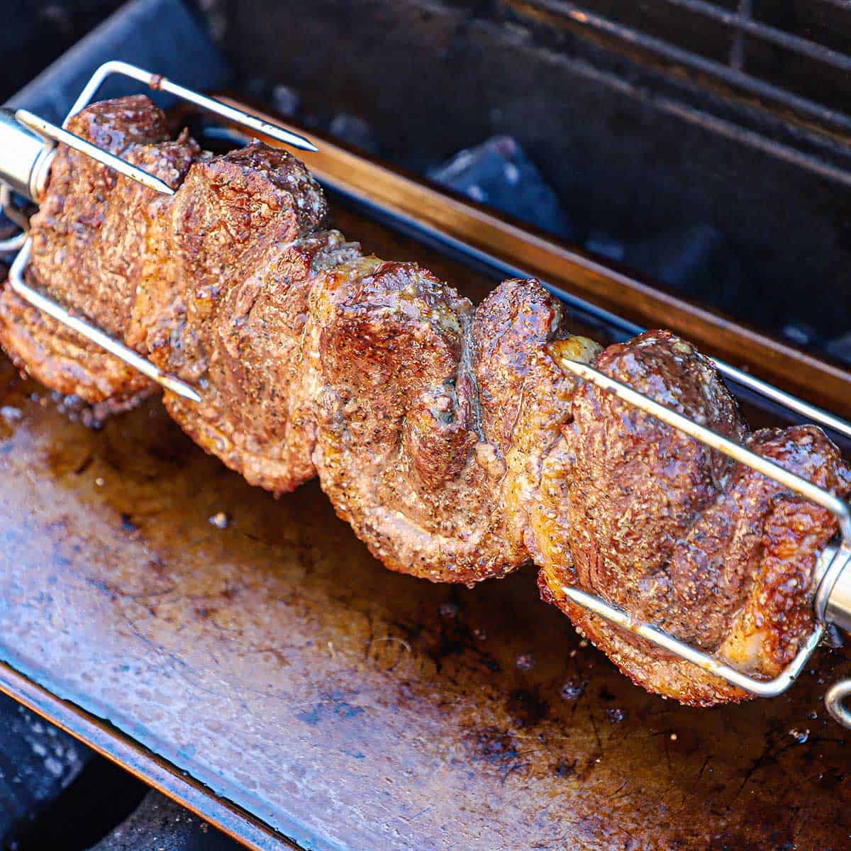 Four folded picanha steaks are being browned and cooked on a rotisserie that is affixed on a gas grill with a metal drip pan underneath the meat.