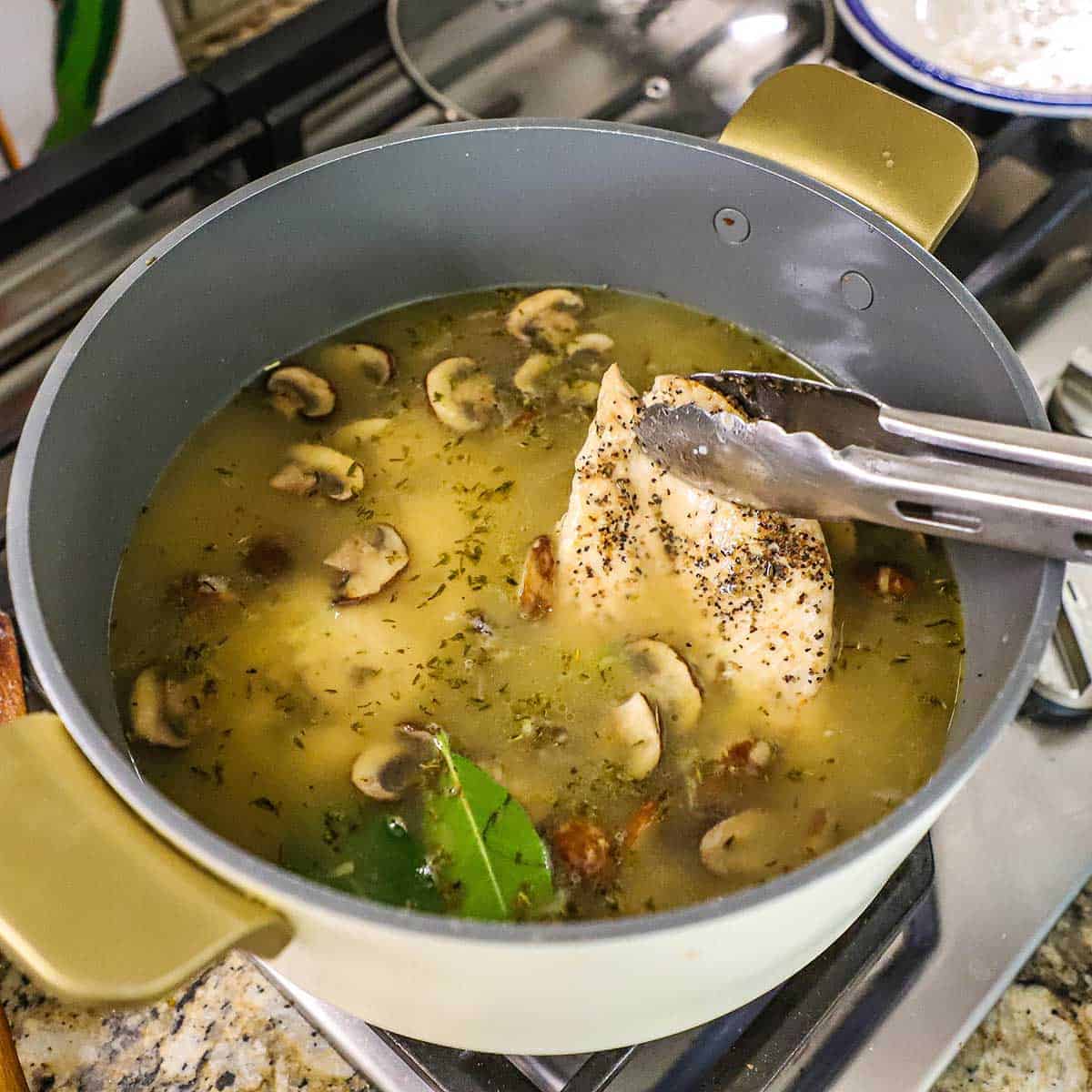 A person placing a seared seasoned chicken breast into a pot filled with simmering chicken broth along with sautéd vegetables, spices, and herbs.