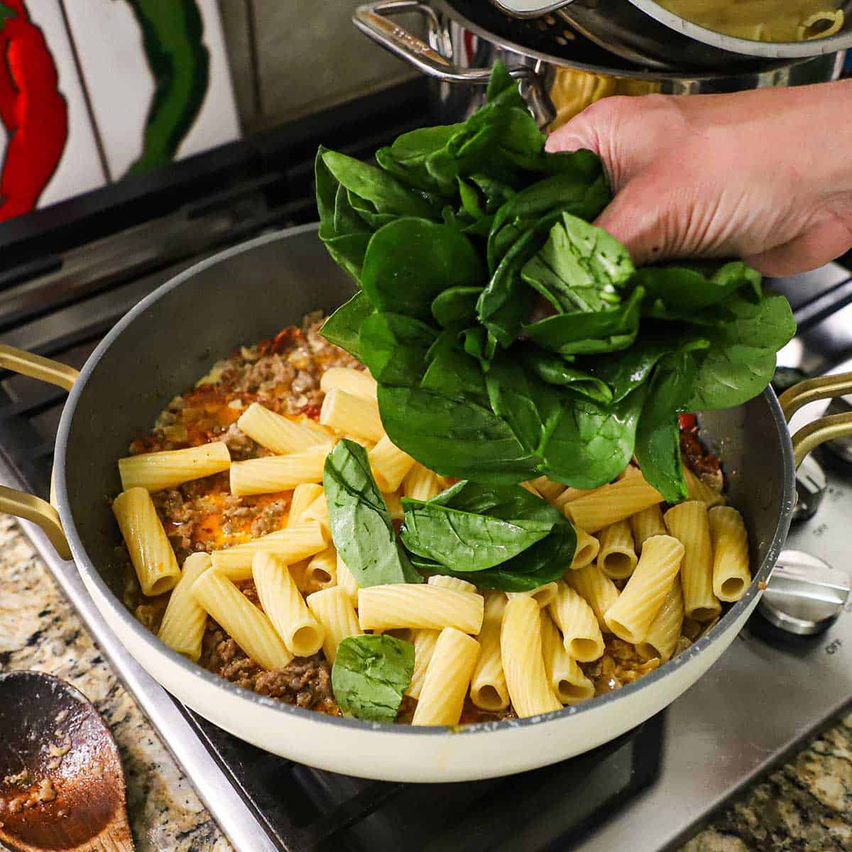 A person using his hand to place a handful of fresh spinach into a large skillet filled with cooked rigatoni pasta and a Tuscan sausage sauce.