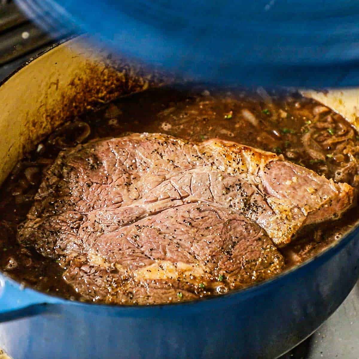 A person placing the lid of a large oval Dutch oven onto the pot that is filled with a simmering French onion sauce with a seared chuck roast in the middle of it all.