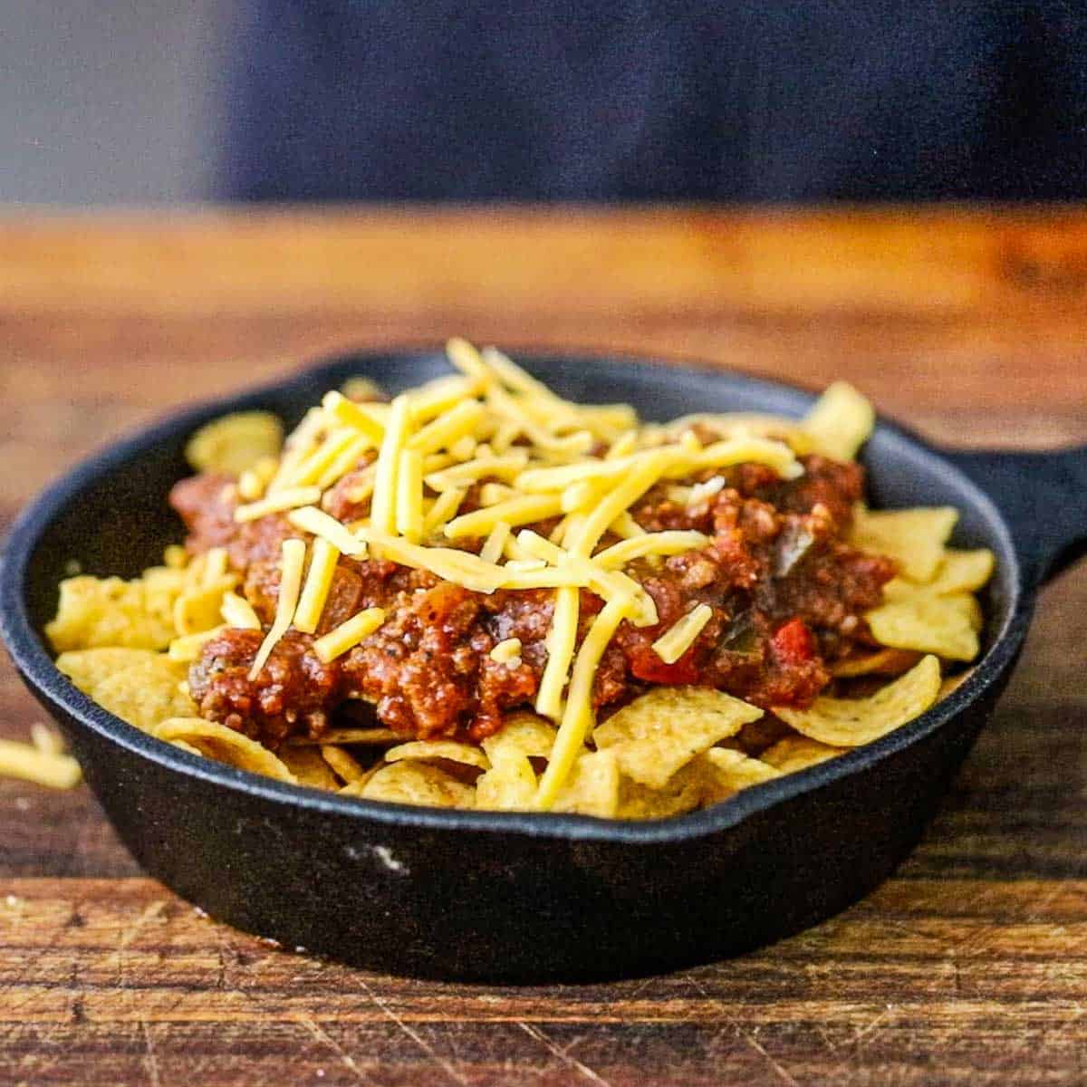A small cast-iron skillet on a wooden cutting board filled with corn chips, topped with beef chili, and shredded cheddar.