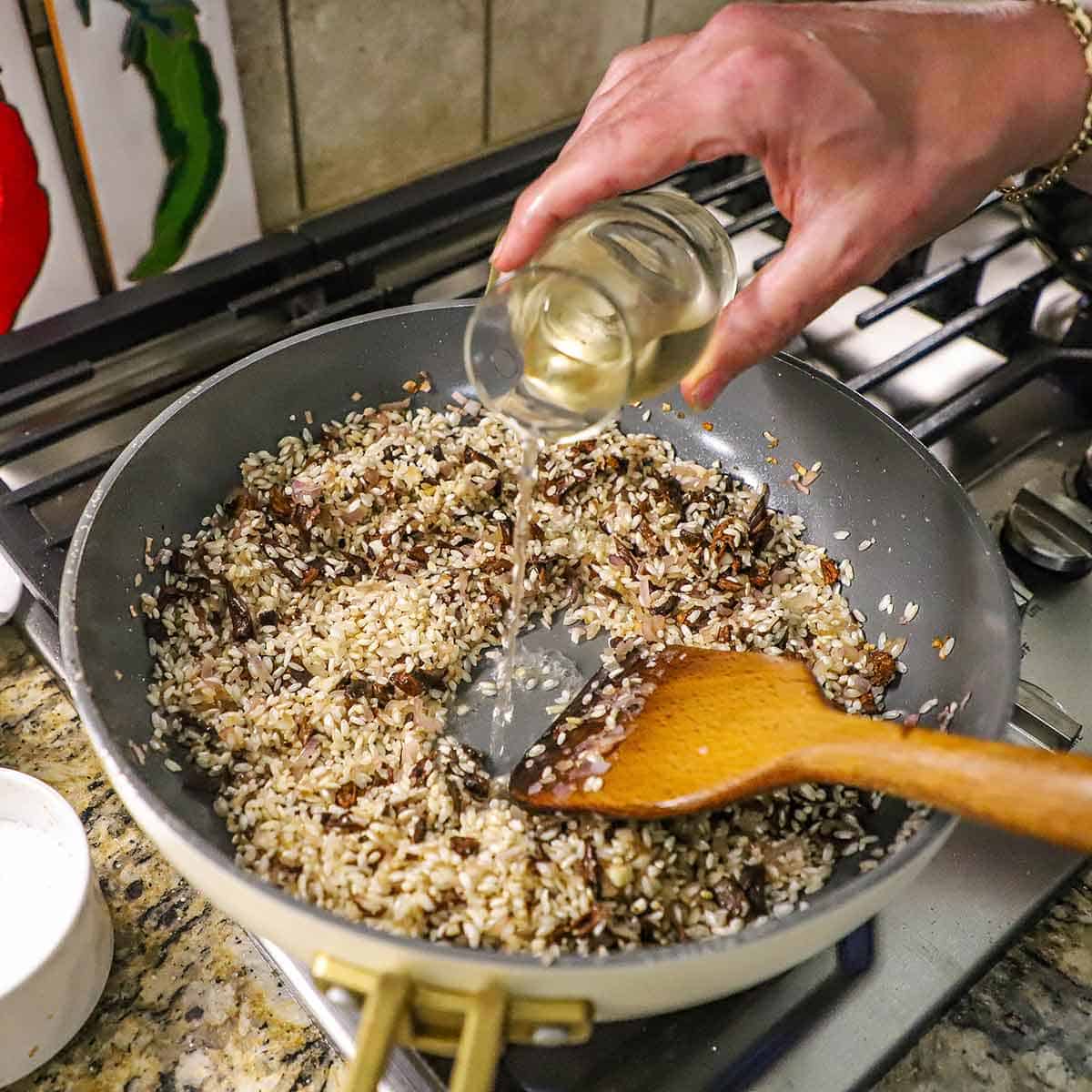 A person pouring white wine from a small carafe into a large skillet filled with toasted arborio rice and saut&eacute;ed shallots and chopped porcini mushrooms.