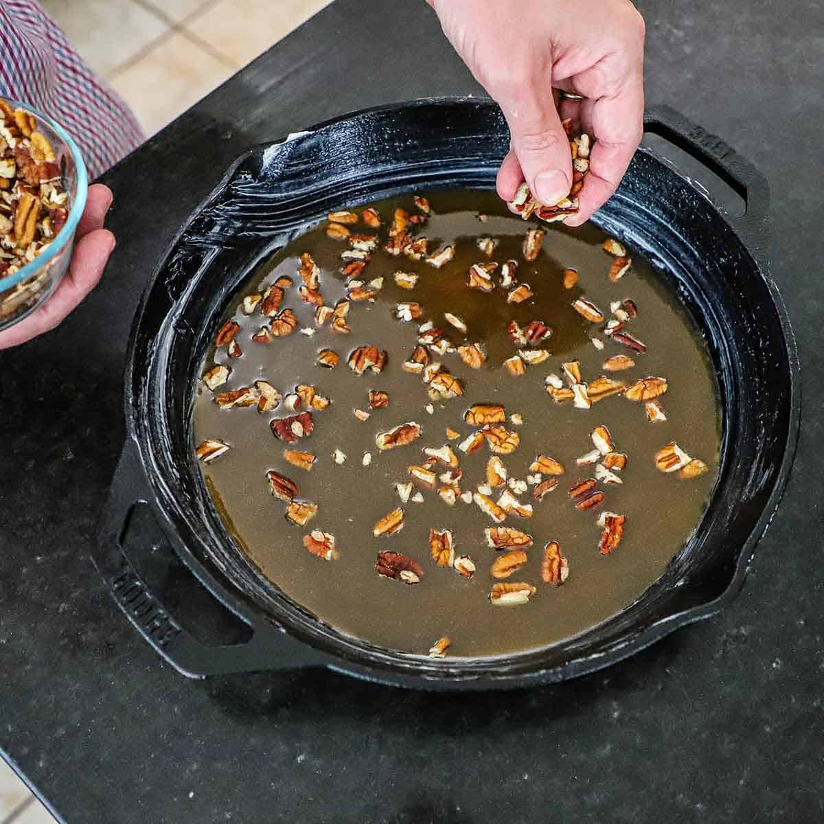 A person sprinkling chopped pecans over the surface of caramel sauce that is resting on the bottom of a large cast-iron skillet.