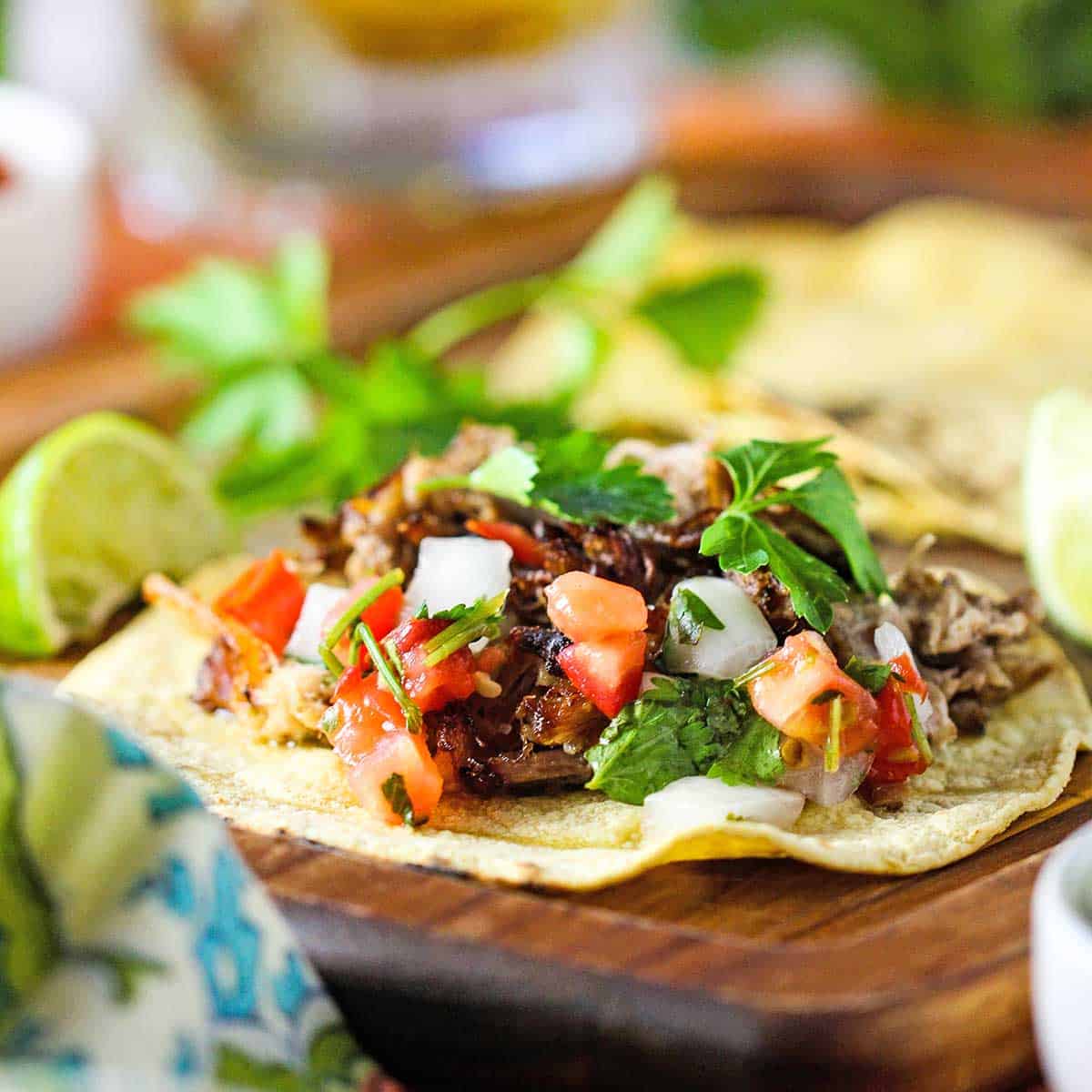 A straight-on view of a flat slow cooker carnitas taco resting open-faced on a cutting board garnished with pico de gallo and cilantro leaves.