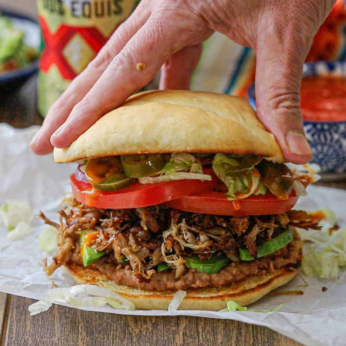A person placing the top portion of a toasted telera bun on the top of the fully loaded carnitas torta packed with beans, pork, vegetables, and crumbled cheese.