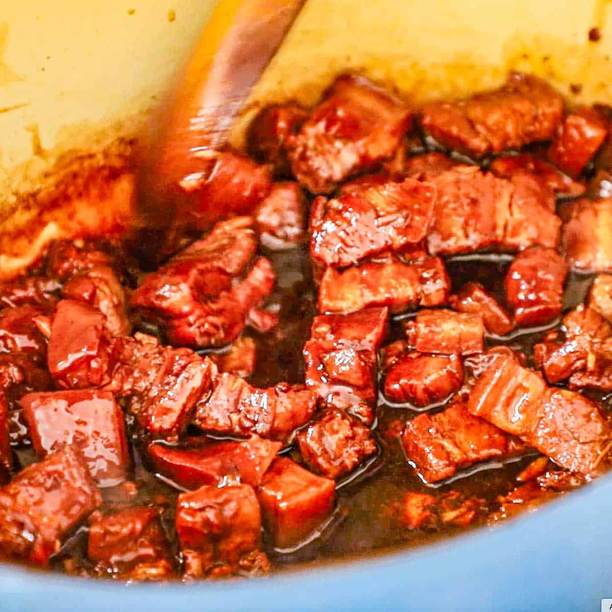 A close-up view of a person using a wooden spoon to stir braised pork belly chunks in a glossy Asian brown sauce inside a large Dutch oven.