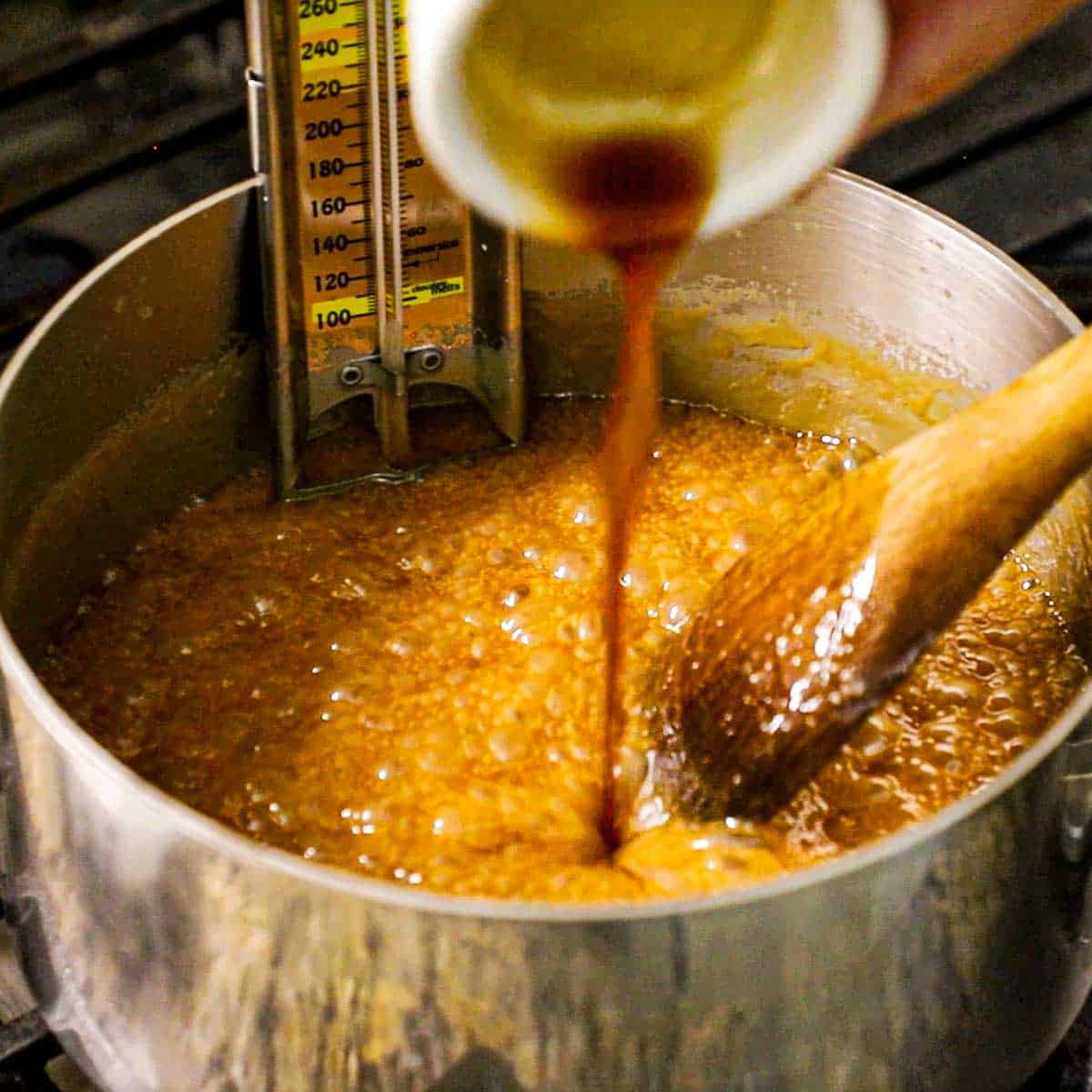 A person pouring vanilla extract from a small white bowl into a saucepan that is filled with hot, bubbling caramel sauce with a candy thermometer attached to the side of the pan.
