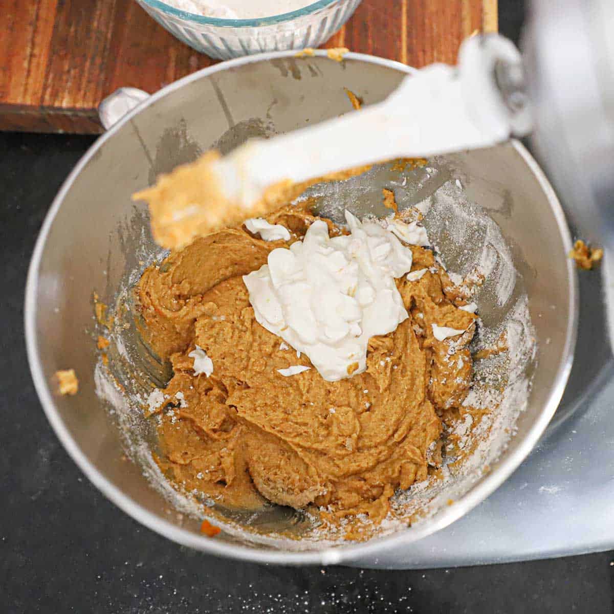 An overhead view of a mound of sour cream that is resting on a pumpkin coffee cake batter in the bowl of a stand mixer with a glass bowl of spiced flour sitting near the mixer.