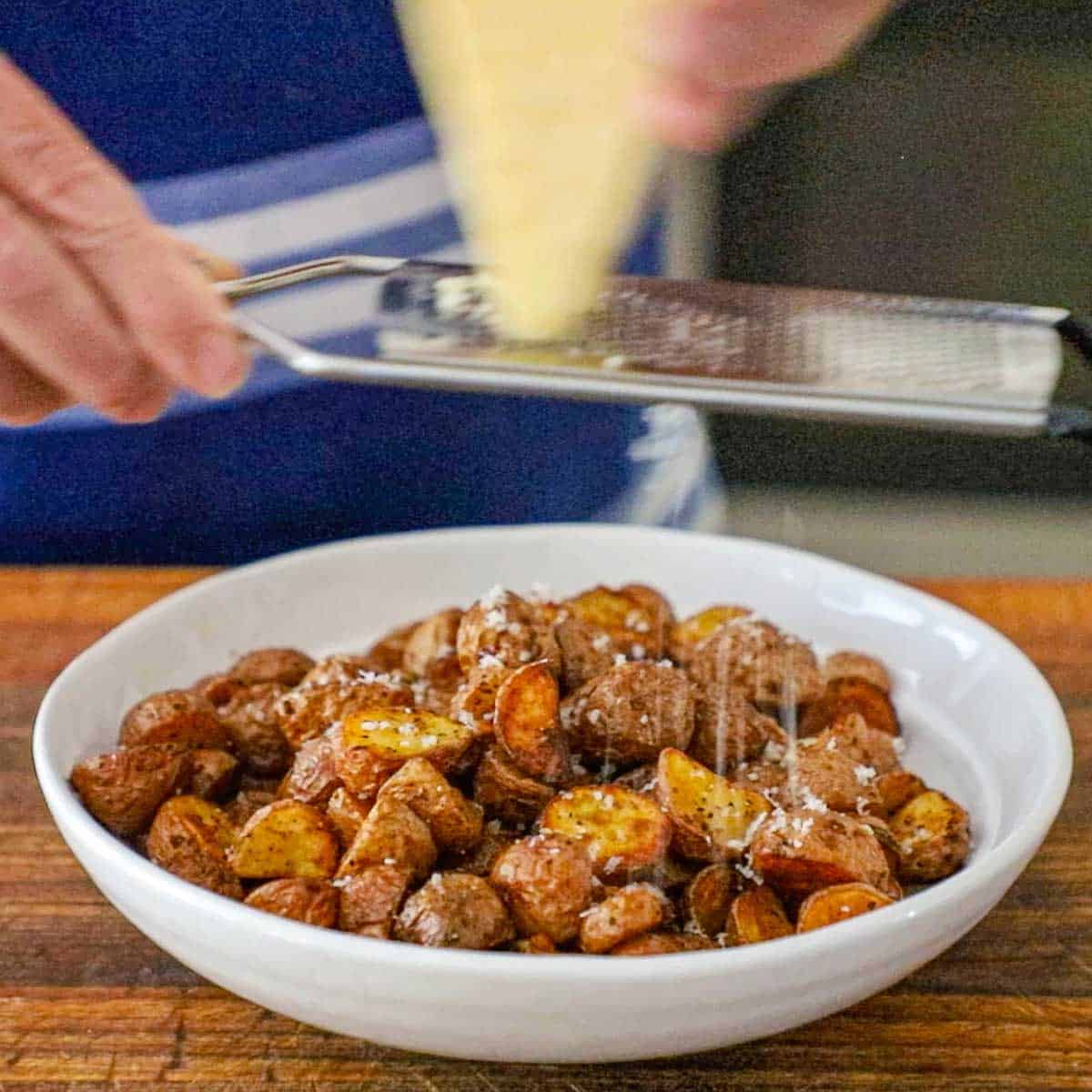 A person grating a block of Parmesan cheese with a microplane over the top of a white shallow bowl filled with a mound of Crispy Air-Fryer Baby Potatoes.
