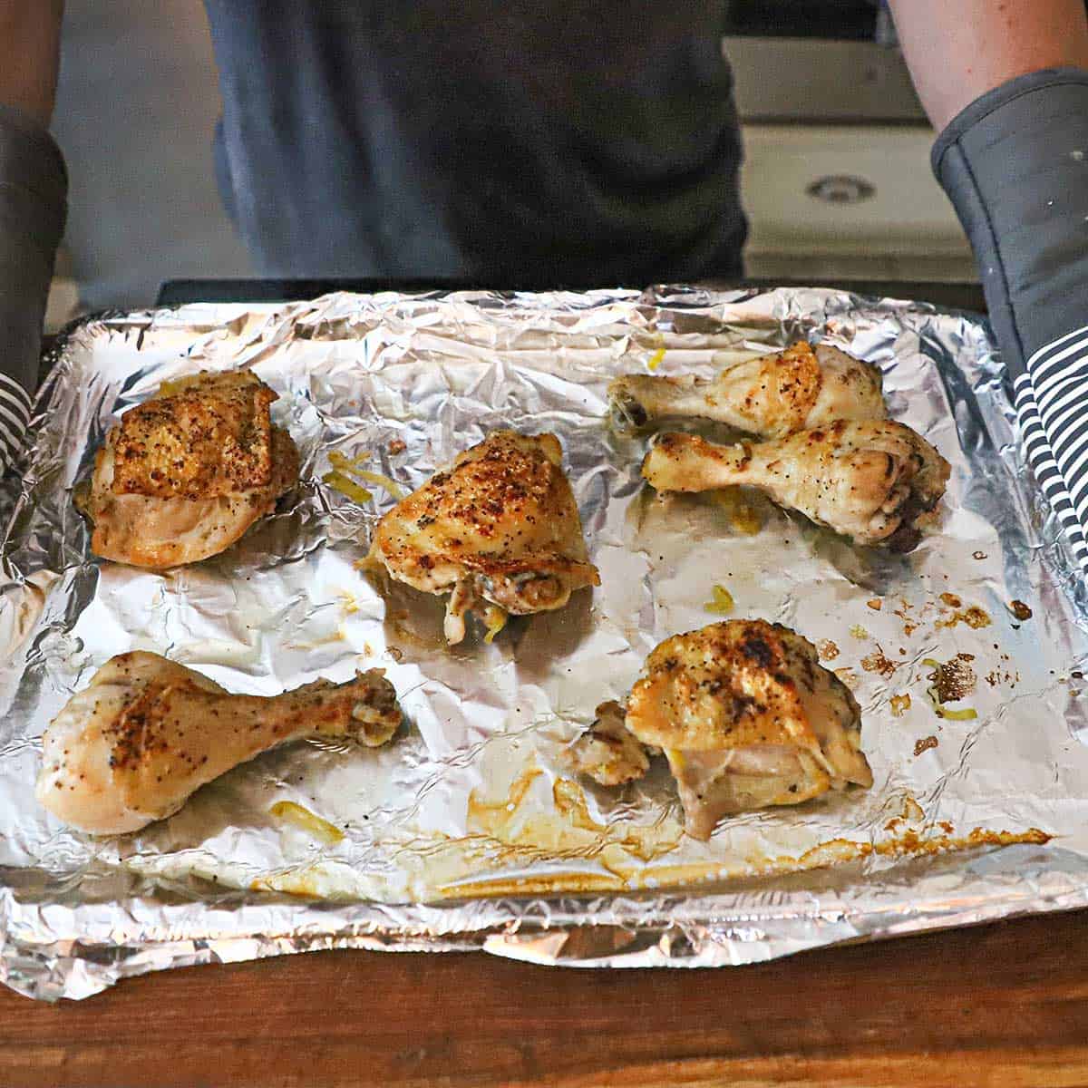 A person using oven mitts to hold a foil-lined baking pan that is holding cooked chicken piece that have just out from under the broiler.