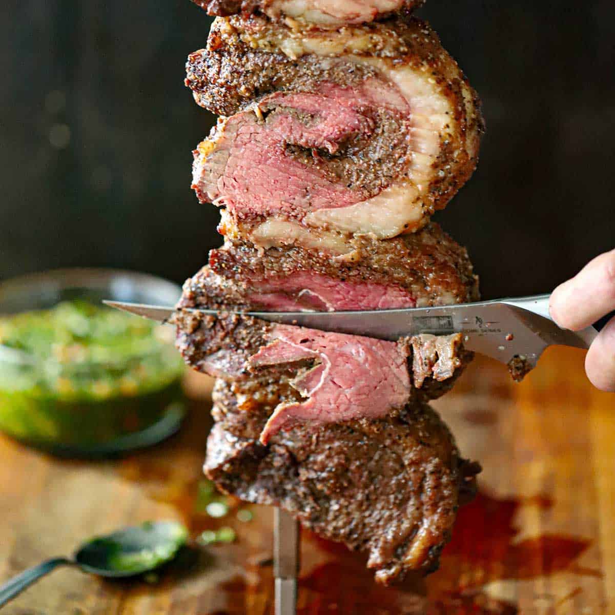 A person using a sharp carving knife to slice thin layers of picanha steak from a one of three folded steaks that are all on a skewer and freshly baked with a rotisserie.