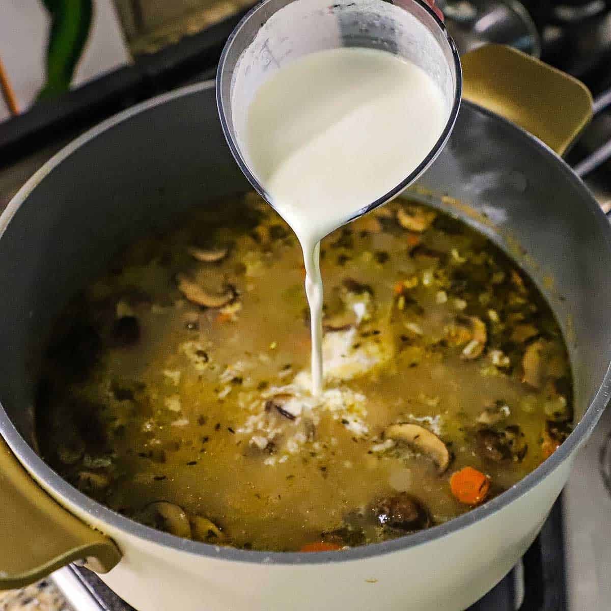A person pouring heavy cream from a plastic measuring cup into a pot of simmering chicken and wild rice soup on a gas stove.