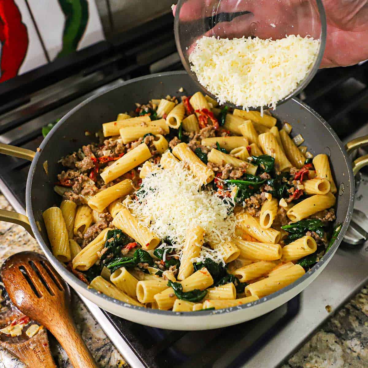 A person transferring grated Parmesan cheese from a glass bowl into a large skillet filled with simmering Tuscan Sausage Pasta on a gas stove.
