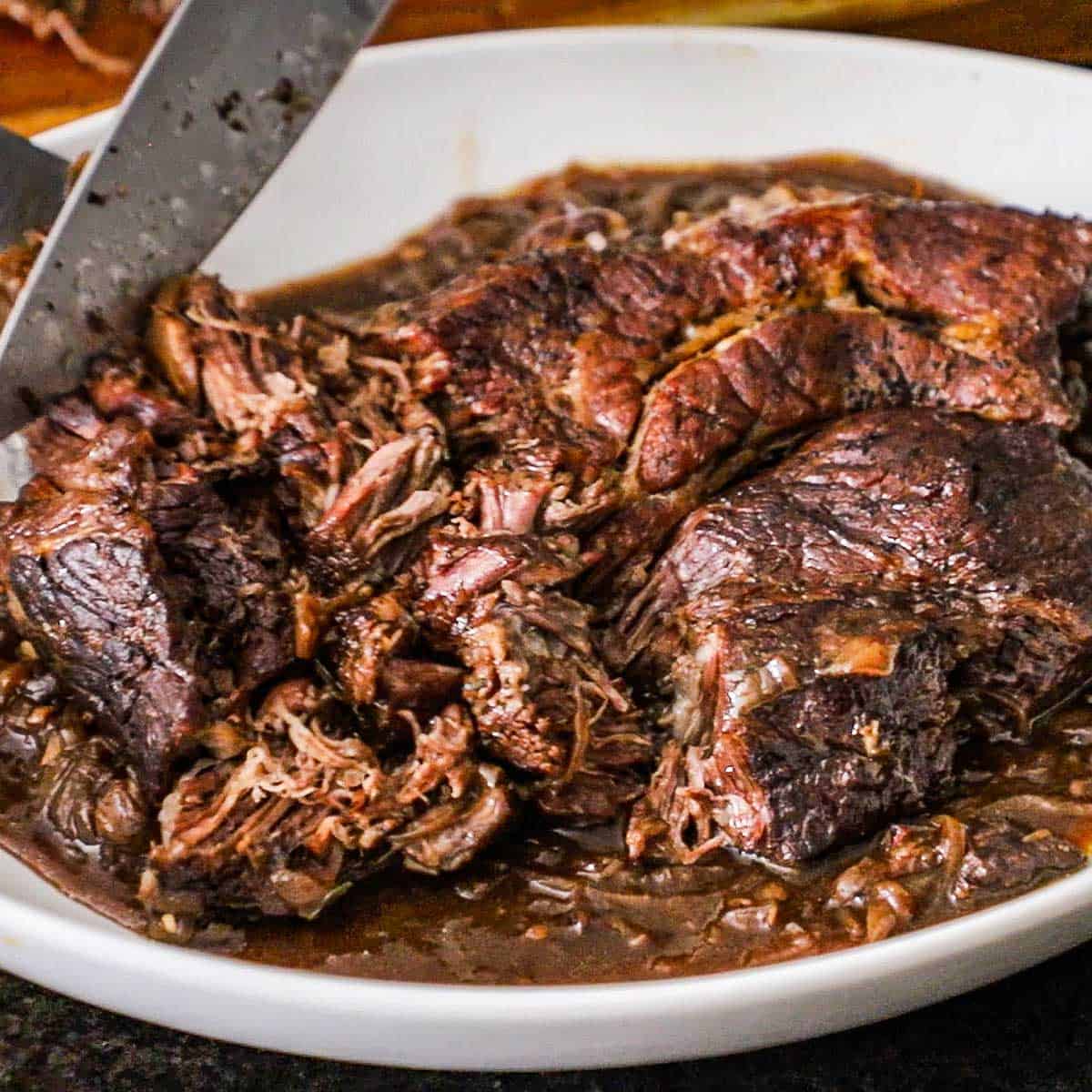 A person using a chef's knife to slice an extremely tender French onion pot roast that is resting along with a brown sauce in a large deep platter.