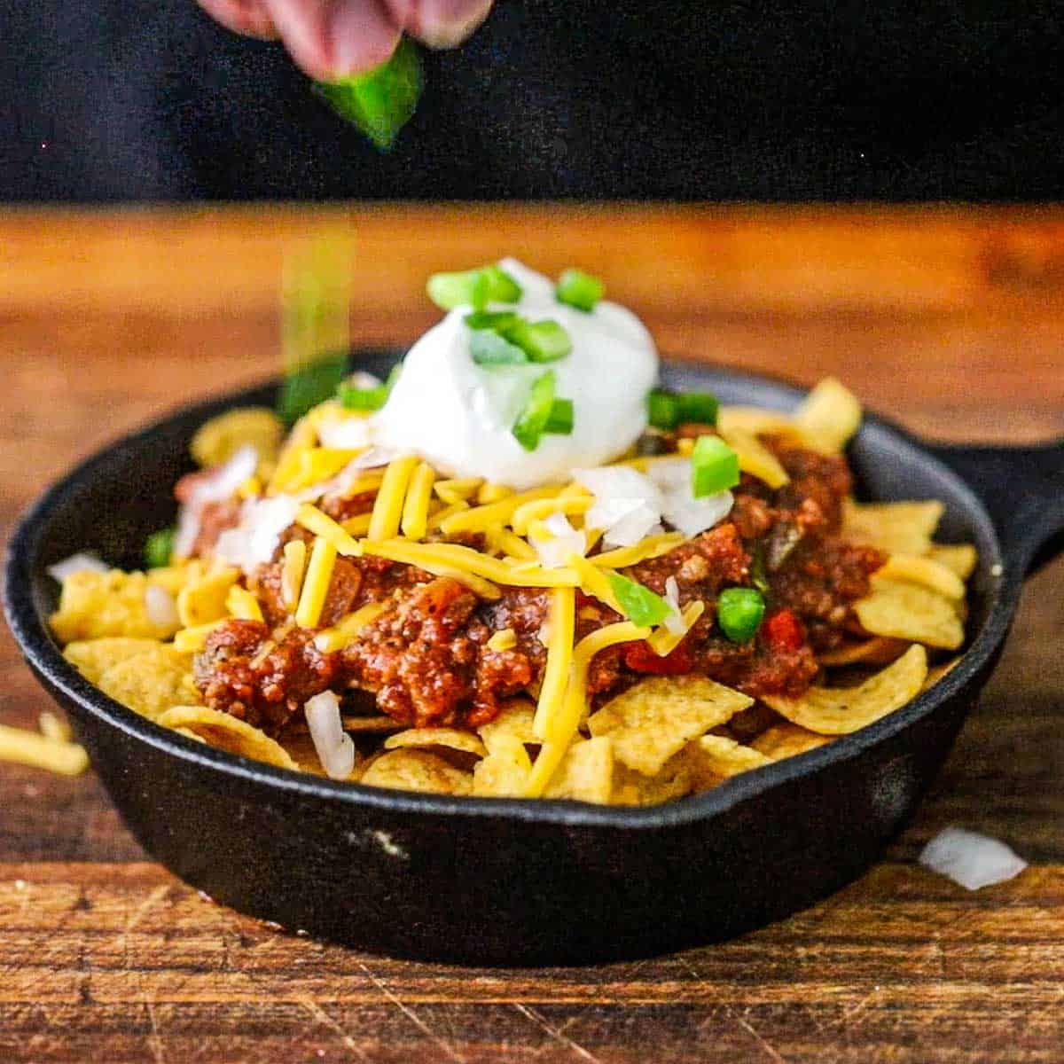 A person using his fingers to garnish a small cast-iron skillet filled with a serving of Classic Frito Pie with chopped jalapeños and a dollop of sour cream.