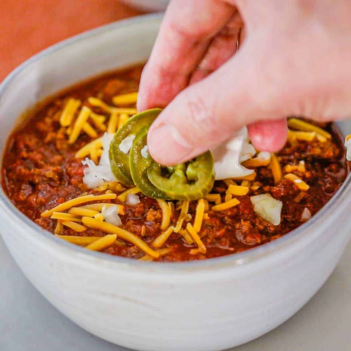 A person placing sliced pickled jalapeños onto the top of a bowl of weeknight beef chili that has also been garnished with shredded cheddar cheese, chopped onions, and a dollop of sour cream.
