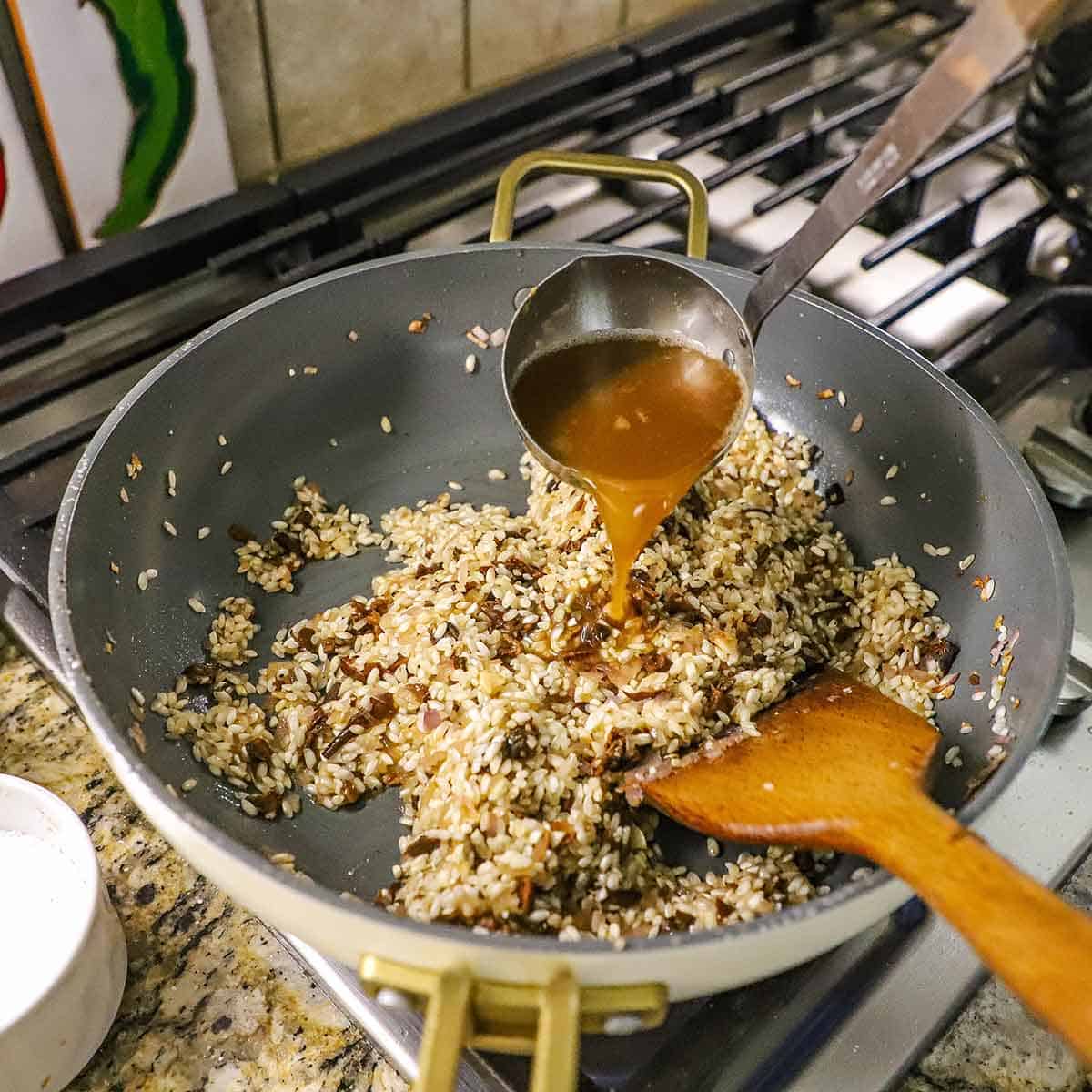 A person using a large metal ladle to transfer hot chicken stock into a skillet filled with toasted arborio rice and saut&eacute;ed shallots and porcini mushrooms.