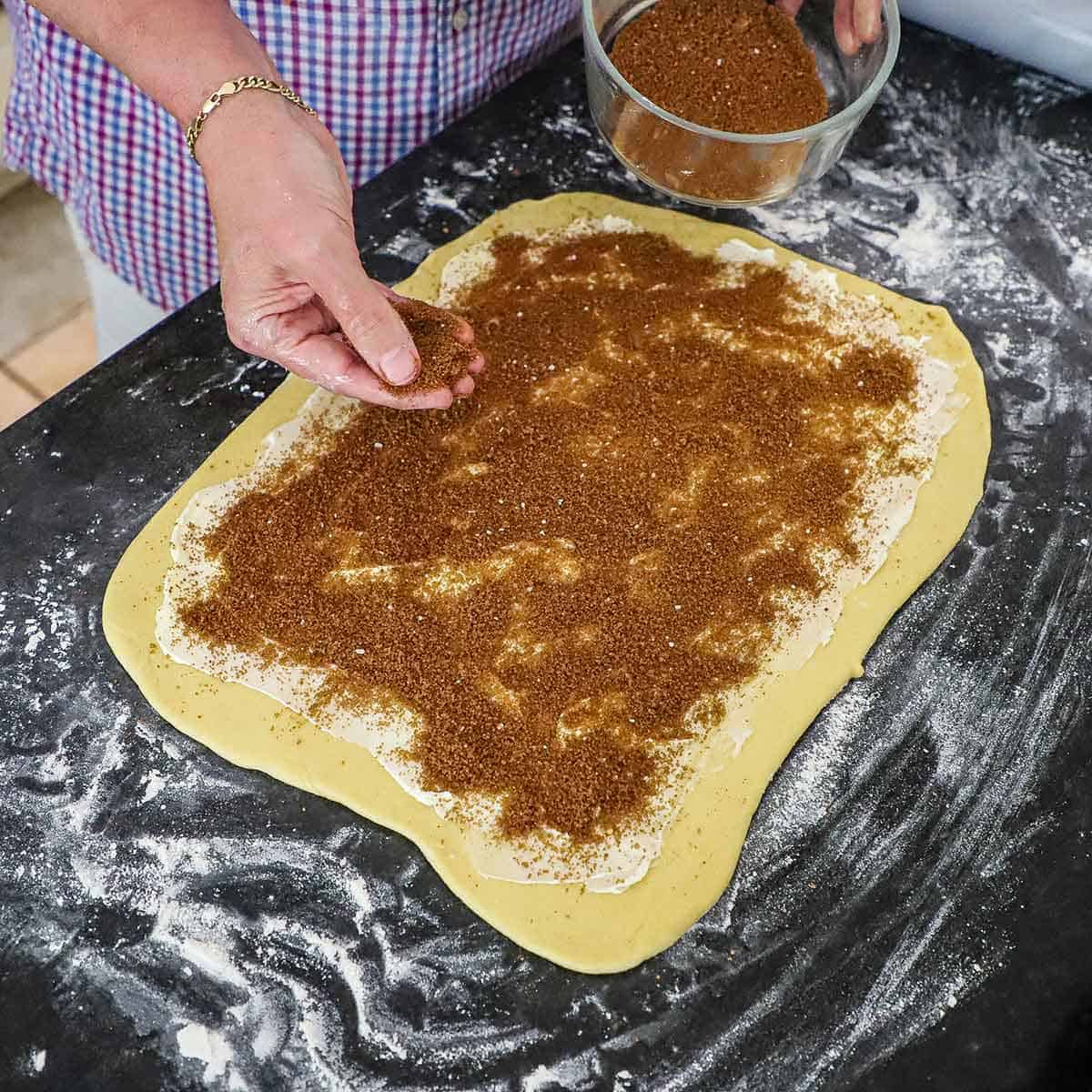 A person sprinkling a brown sugar and cinnamon mixture over a large rectangle of cinnamon roll dough that is on a lightly floured surface and has a layer of softened butter between the dough and the sugar mixture.