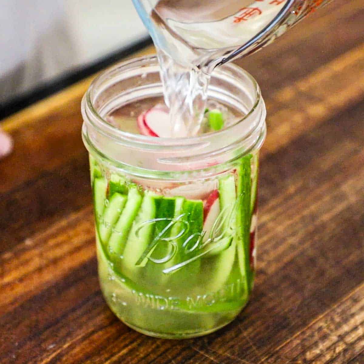 A person pouring warm pickling liquid from a glass measuring cup into a glass jar that is filled with thin strips of cucumber and thin slices of radishes.
