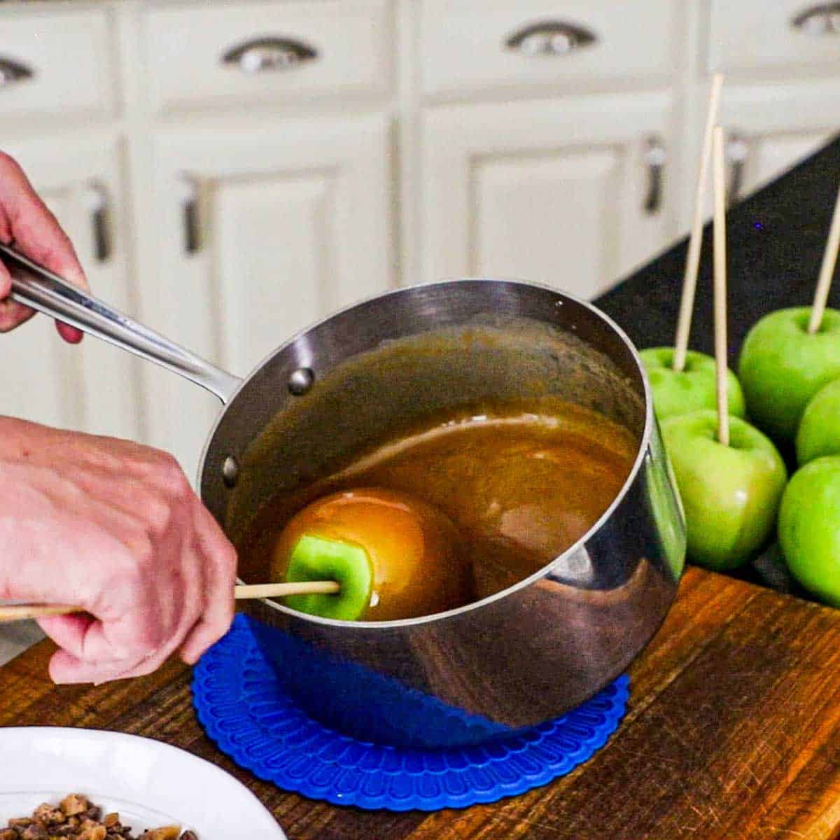 A person tilting a silver saucepan which is causing slightly cooled caramel sauce to pool, allowing hims to turn an apple through the sauce by holding onto the wooden stick that has been inserted into the top of the apple.