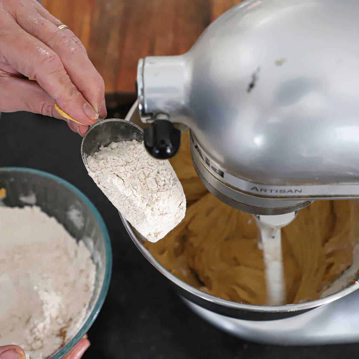 A person using a scooper to transfer spiced flour into the bowl of a stand mixer that is mixing a pumpkin spice coffee cake batter.