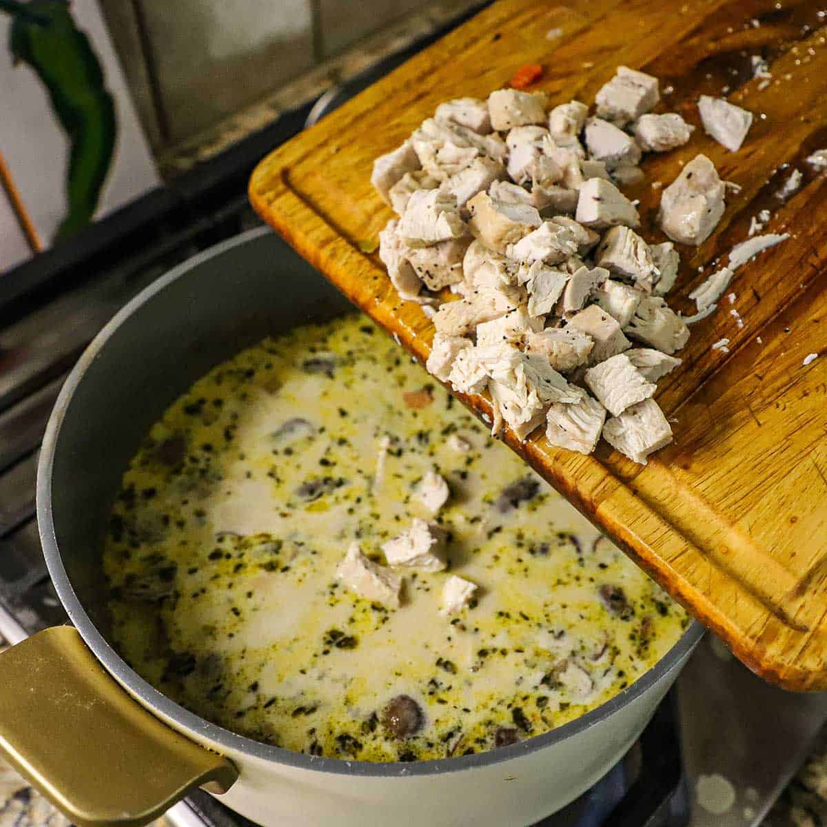 A person transferring cubed, cooked chicken from a wooden cutting board into a pot filled with a simmering wild rice soup.