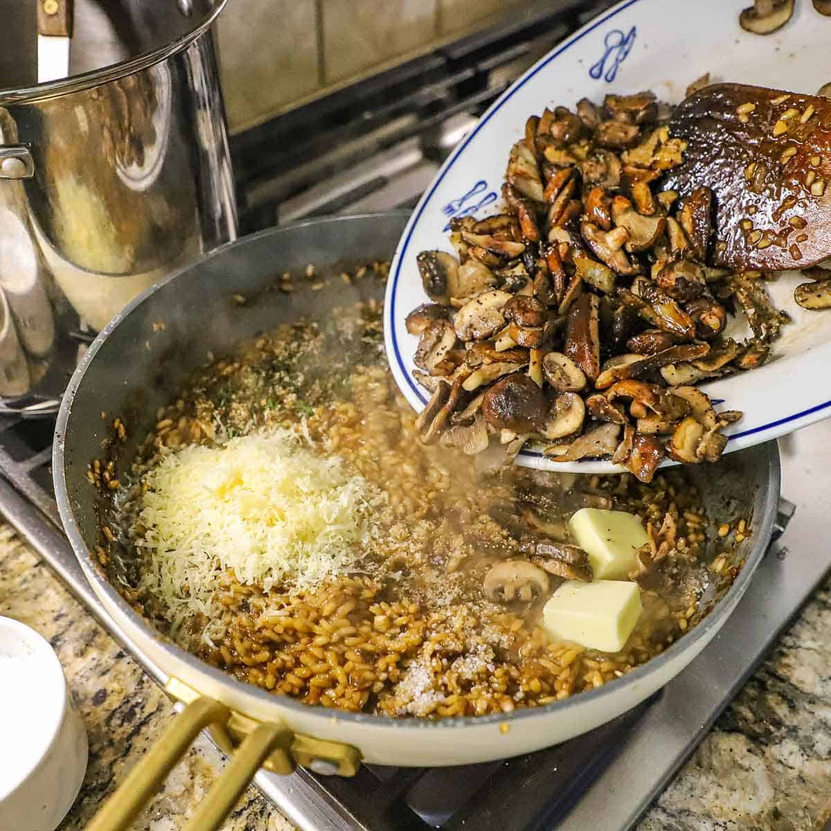 A person transferring saut&eacute;ed sliced wild mushrooms into a skillet filled with simmering wild mushroom risotto including shredded Parmesan cheese and two pallets of unsalted butter.