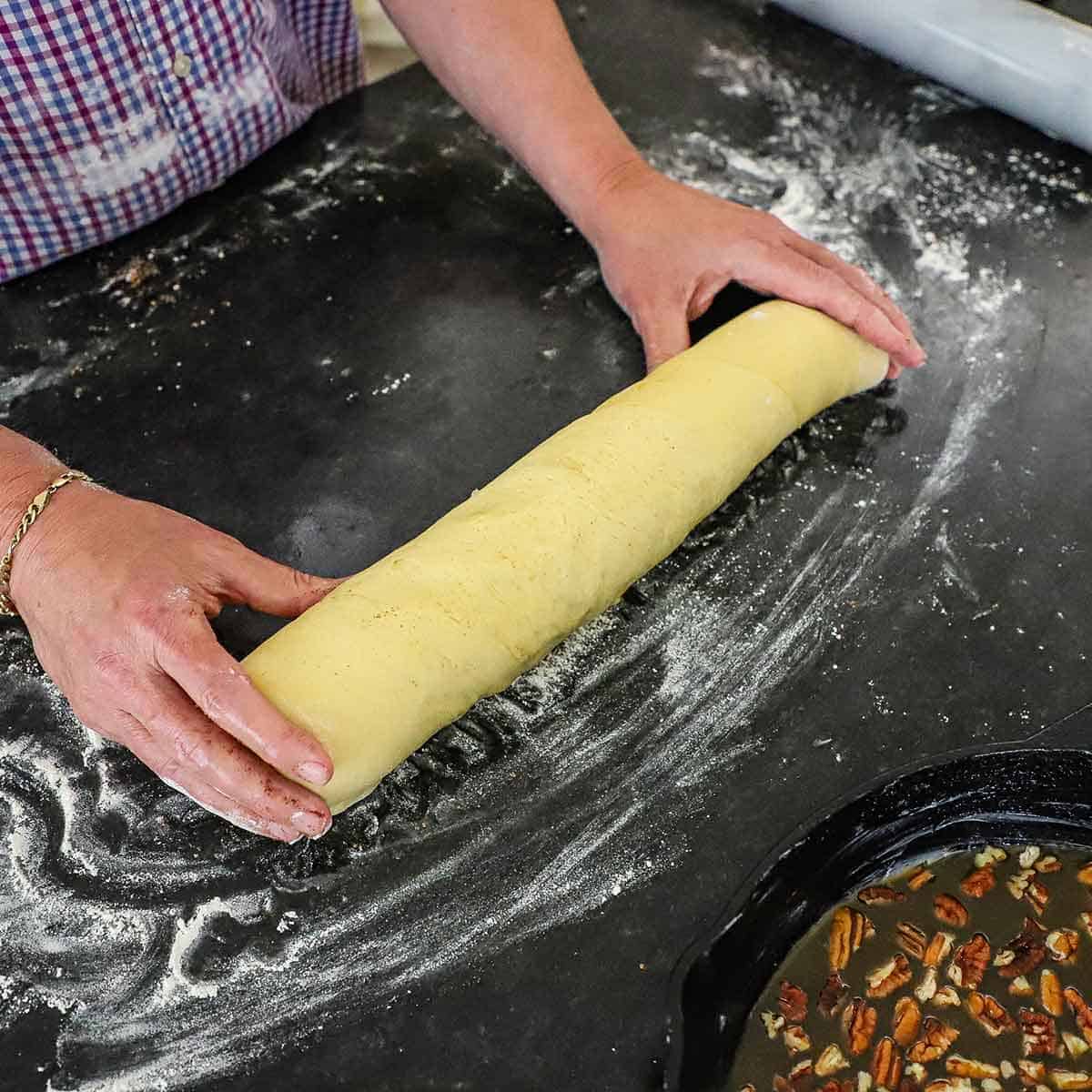 A person holding the ends of a long tube of cinnamon roll dough that has been rolled tightly onto a lightly floured black marble surface.