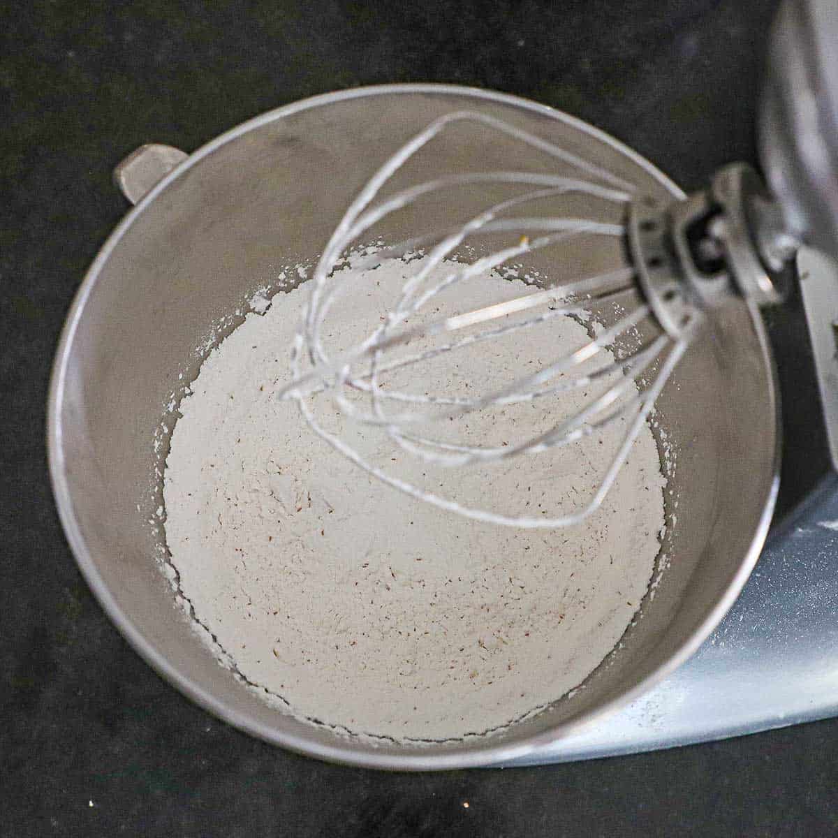 An overhead view of a stand-mixer with the whisk attachment sitting over the bowl of combined flour with yeast, sugar, salt, and baking powder.